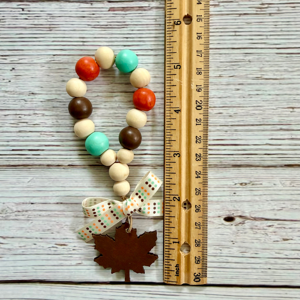 A beaded garland with orange, teal, brown, and cream wooden beads, a patterned ribbon bow, and a small maple leaf charm, displayed next to a wooden ruler on a whitewashed wood background.