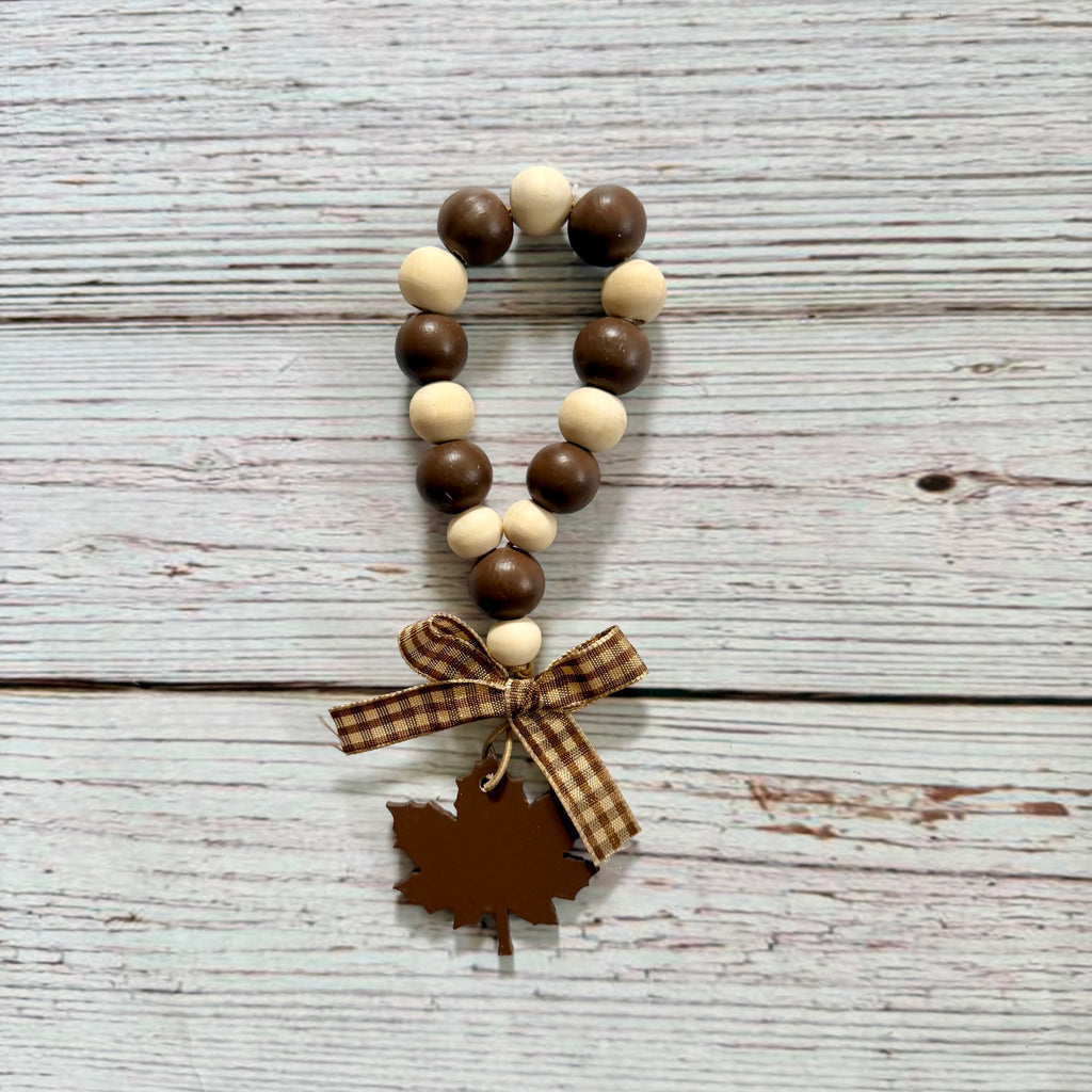A wooden bead garland with alternating dark brown and light cream beads, tied with a brown plaid ribbon and finished with a brown leaf-shaped charm, displayed on a rustic white-washed wood background.