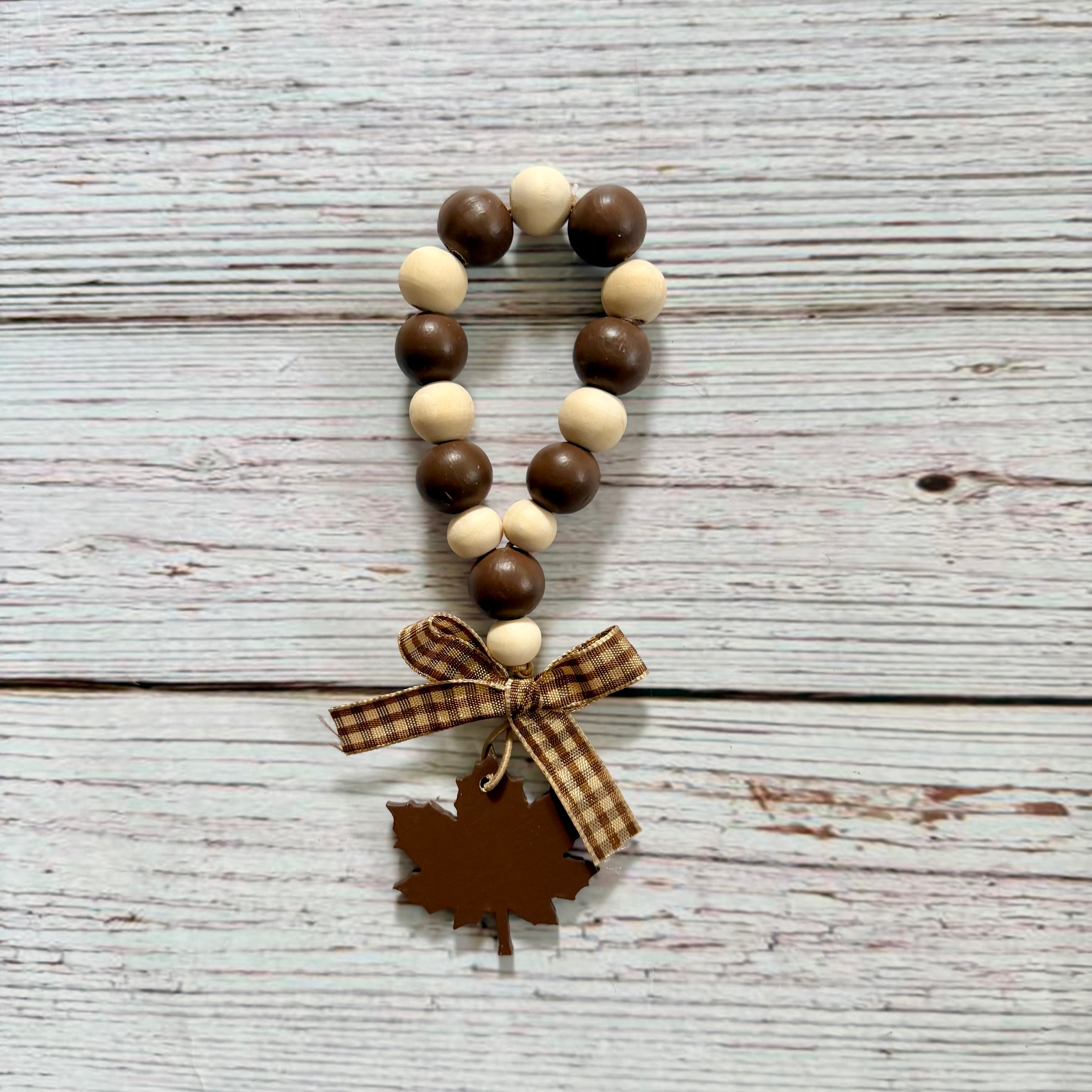 A wooden bead garland with alternating dark brown and light cream beads, tied with a brown plaid ribbon and finished with a brown leaf-shaped charm, displayed on a rustic white-washed wood background.
