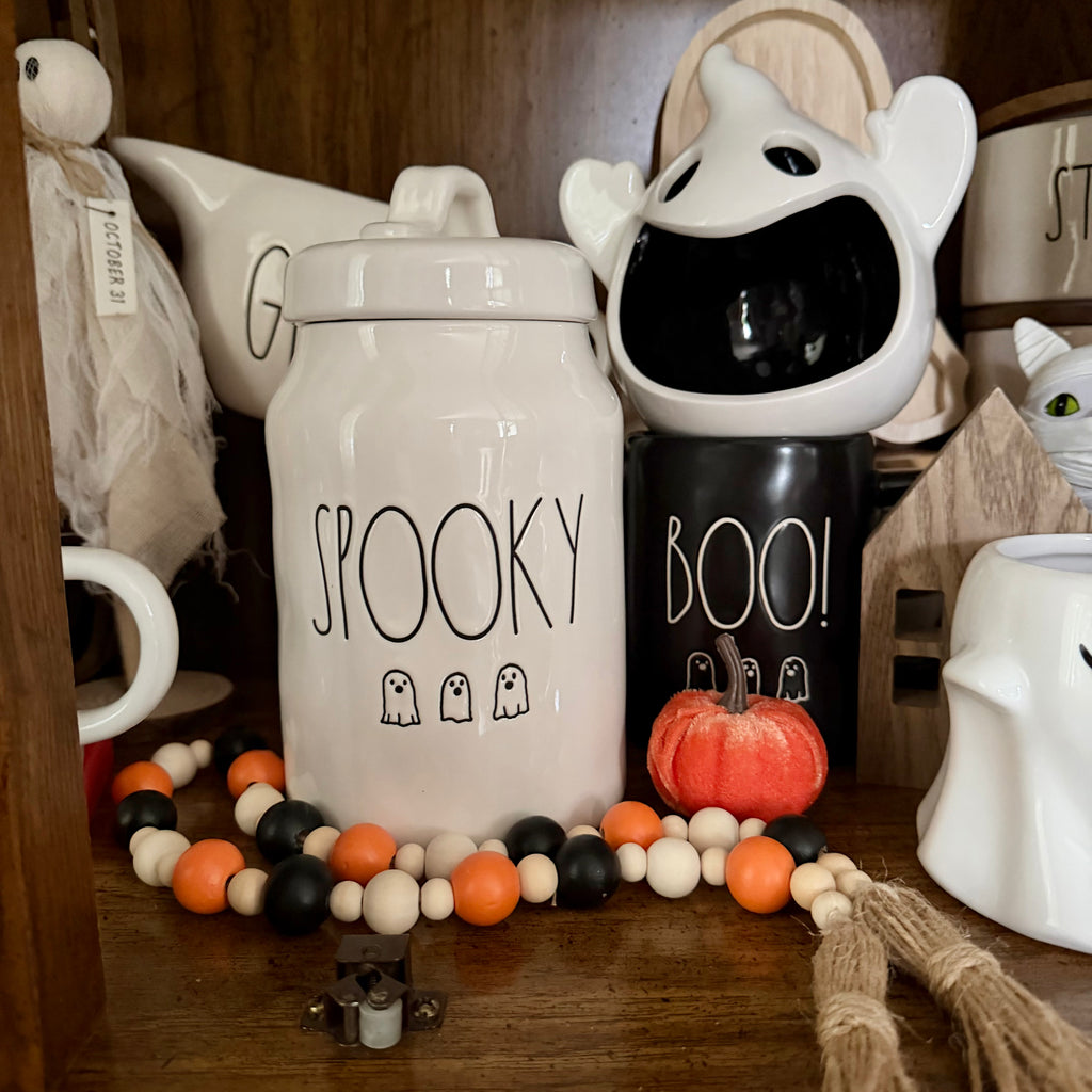 A styled Halloween display featuring ceramic canisters with 'SPOOKY' and 'BOO!' text, a small orange pumpkin, and a beaded garland on a wooden shelf.