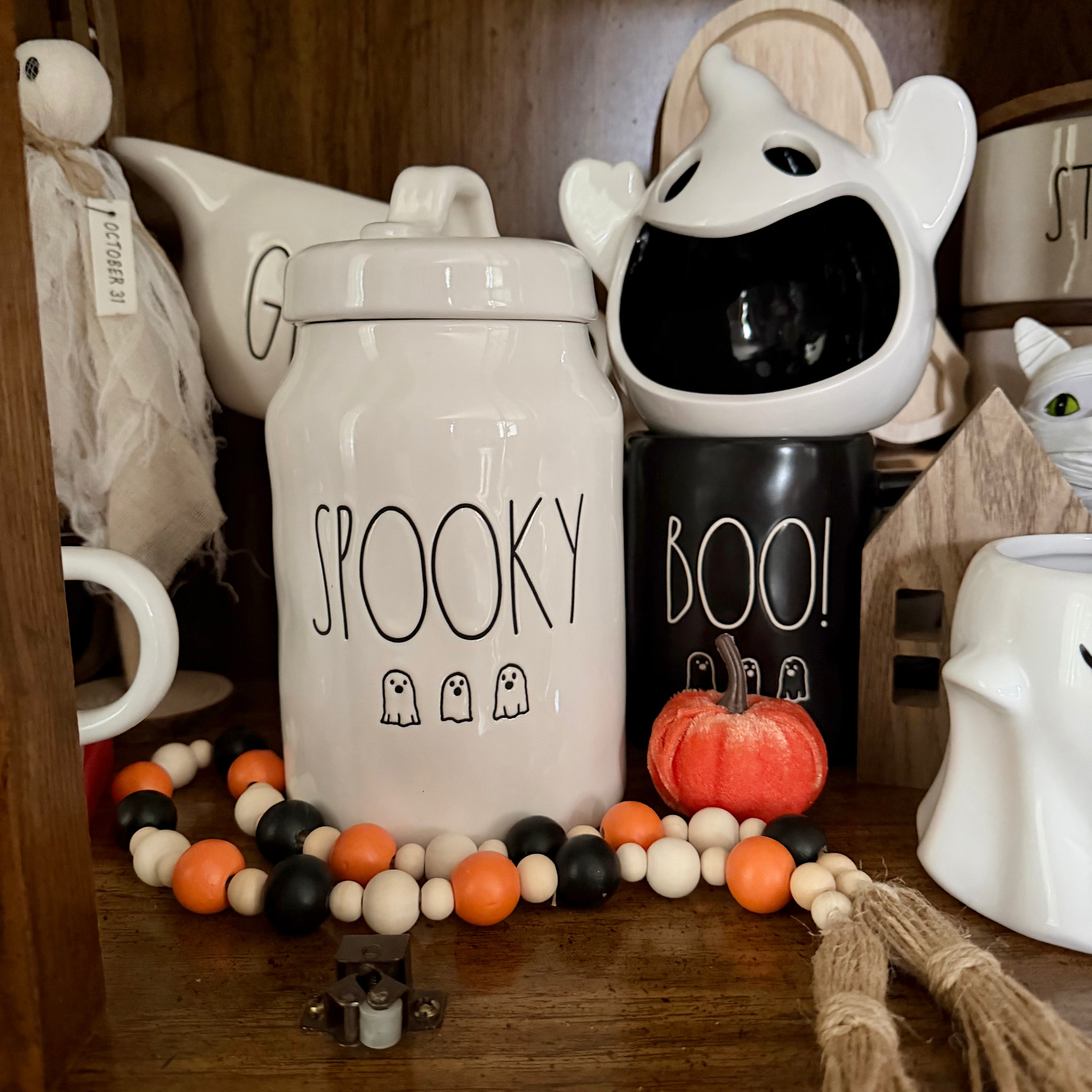 A styled Halloween display featuring ceramic canisters with 'SPOOKY' and 'BOO!' text, a small orange pumpkin, and a beaded garland on a wooden shelf.