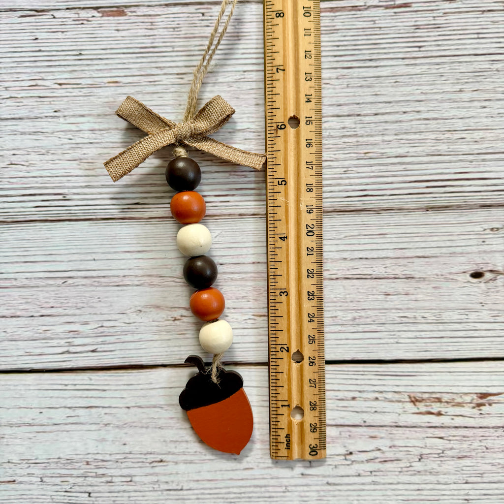 Acorn-shaped beaded hanging ornament with a burlap bow, shown next to a wooden ruler on a whitewashed wood background.