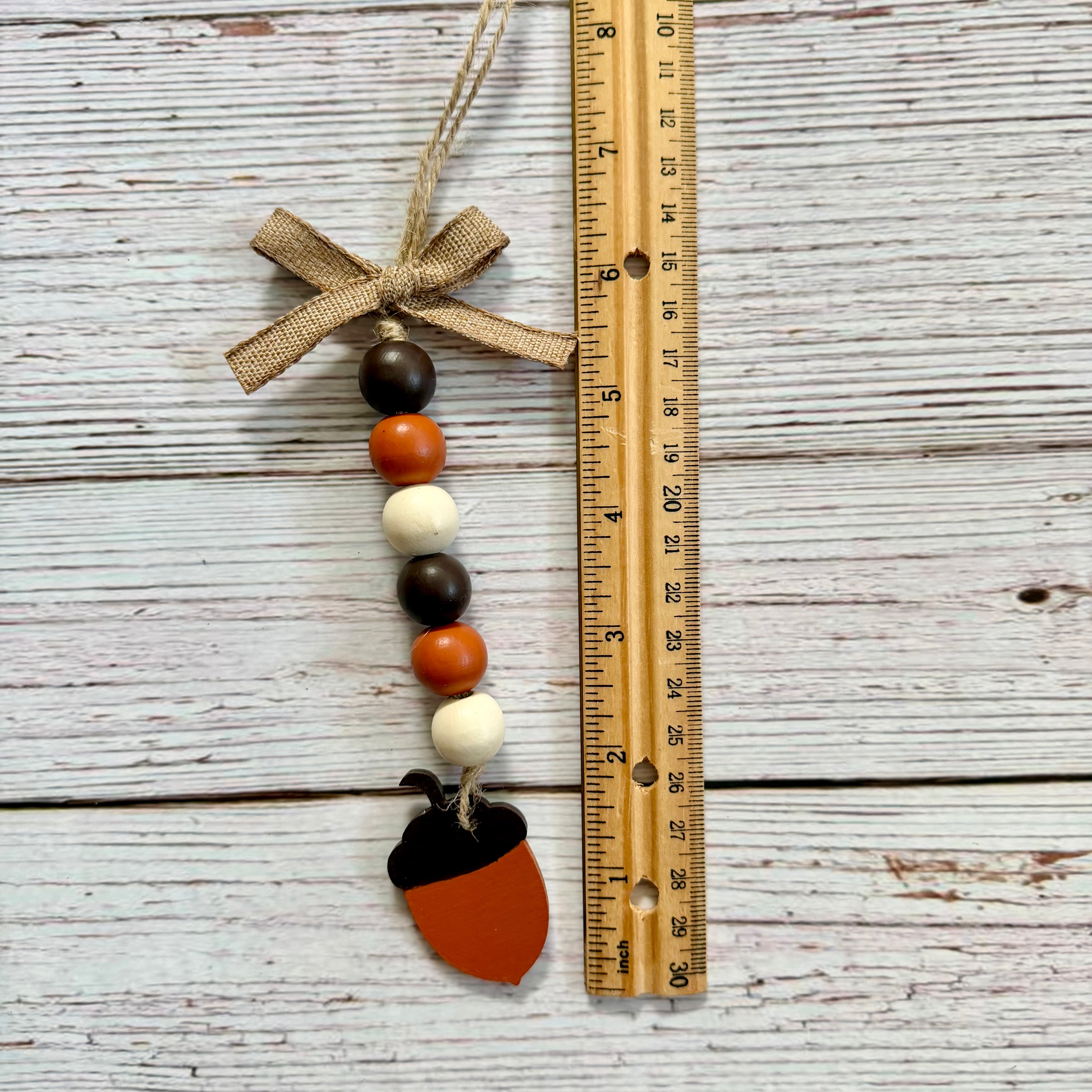 Acorn-shaped beaded hanging ornament with a burlap bow, shown next to a wooden ruler on a whitewashed wood background.