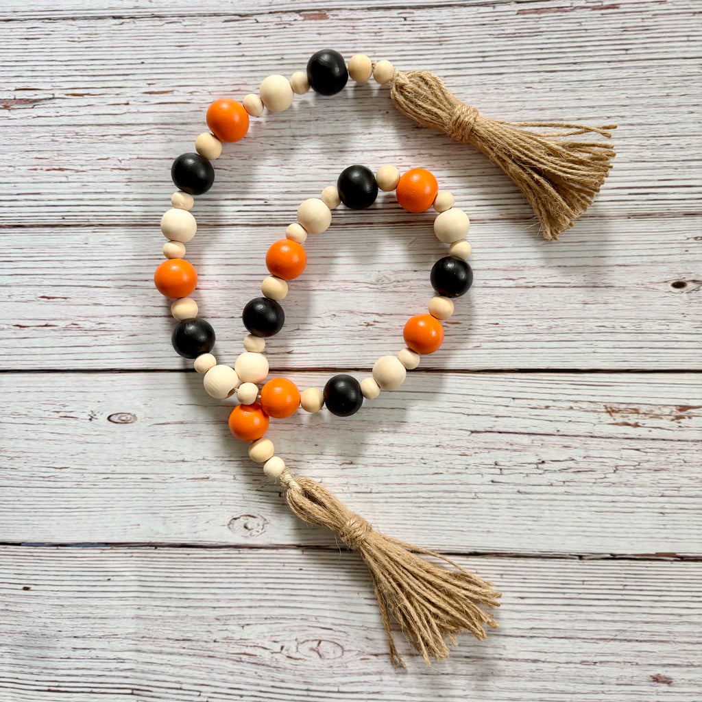 A beaded garland with alternating black and natural wood beads, bright orange accent beads, and two natural jute tassels, displayed on a whitewashed wooden surface.