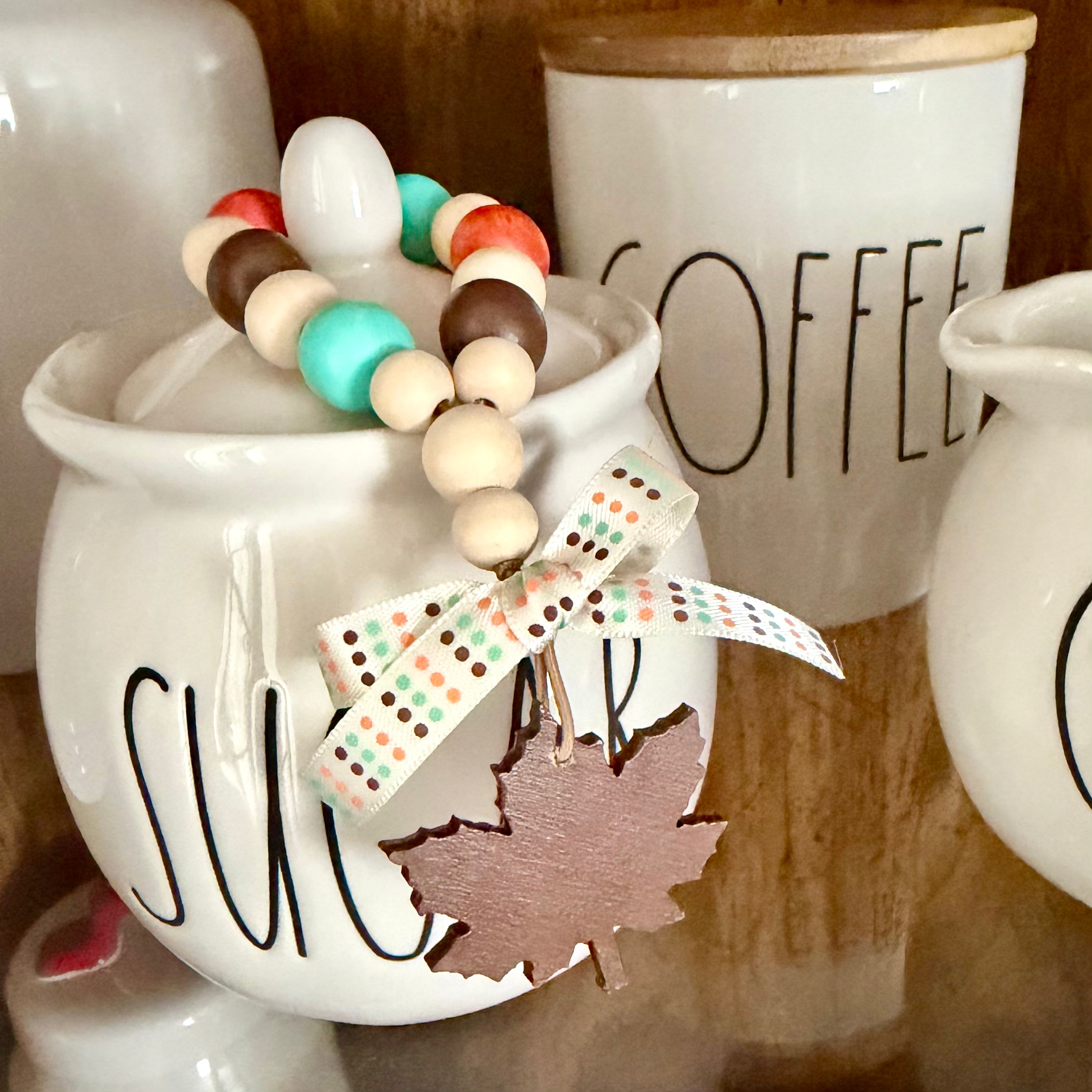 A decorative beaded loop with white, brown, orange, teal, and cream beads, tied with a polka-dot ribbon and a wooden maple leaf charm, draped over a white ceramic sugar canister.