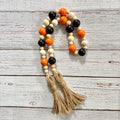 A beaded garland with alternating orange, black, and natural wood beads, finished with two tan jute tassels, displayed on a white-washed wooden background.