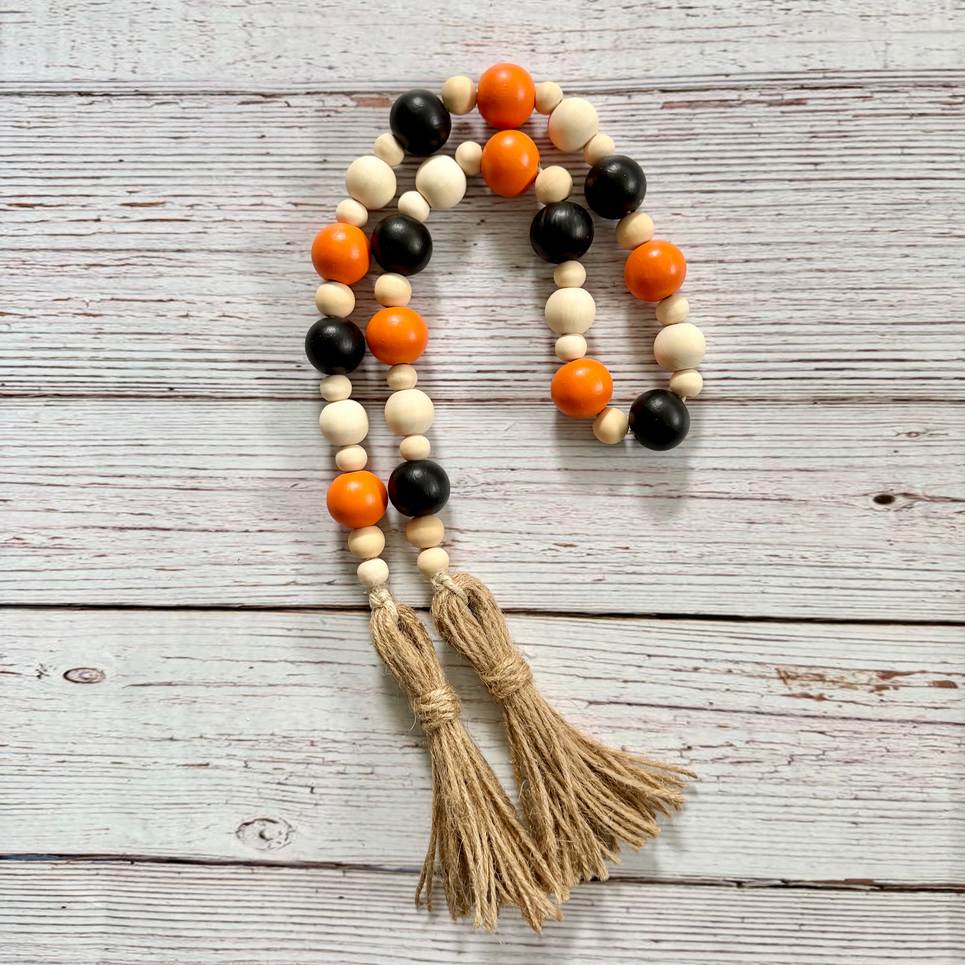 A beaded garland with alternating orange, black, and natural wood beads, finished with two tan jute tassels, displayed on a white-washed wooden background.