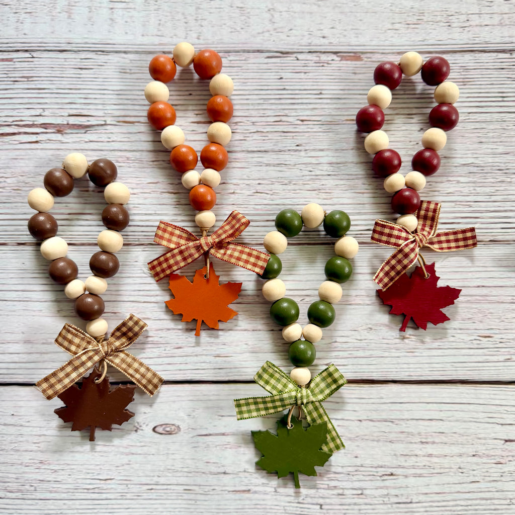 Four autumn-themed beaded keychains with wooden maple leaf charms and gingham ribbon bows, arranged on a white-washed wooden background.