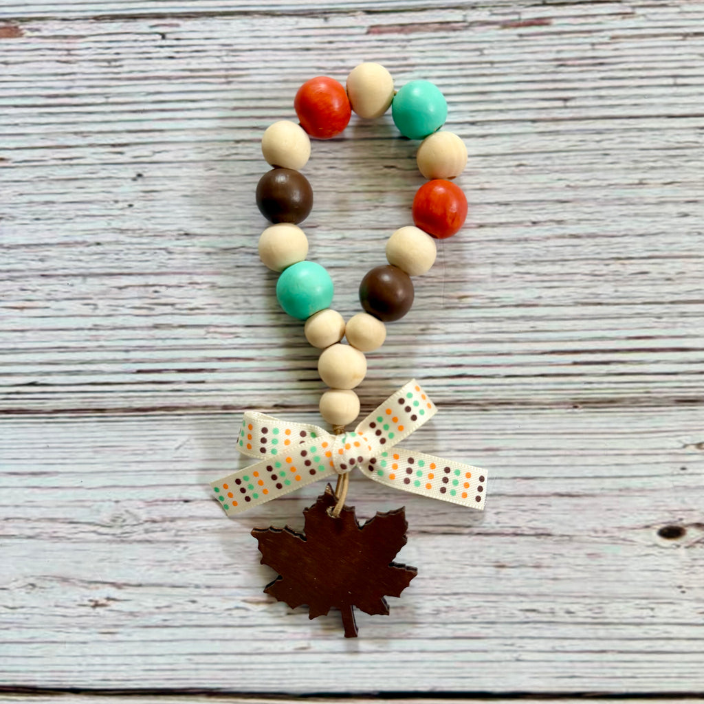 A beaded loop charm with wooden beads in cream, teal, brown, and orange, finished with a cream ribbon bow printed with orange and brown dots and a dark brown wooden maple leaf charm, displayed on a rustic white-washed wood background.