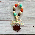 A beaded loop charm with wooden beads in cream, teal, brown, and orange, finished with a cream ribbon bow printed with orange and brown dots and a dark brown wooden maple leaf charm, displayed on a rustic white-washed wood background.