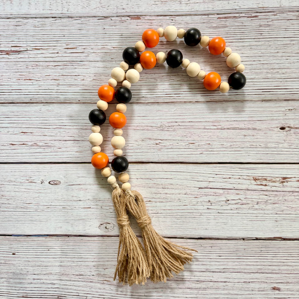 A beaded garland with alternating black, cream, and orange wooden beads, finished with two knotted jute tassels, displayed on a rustic white-washed wooden background.