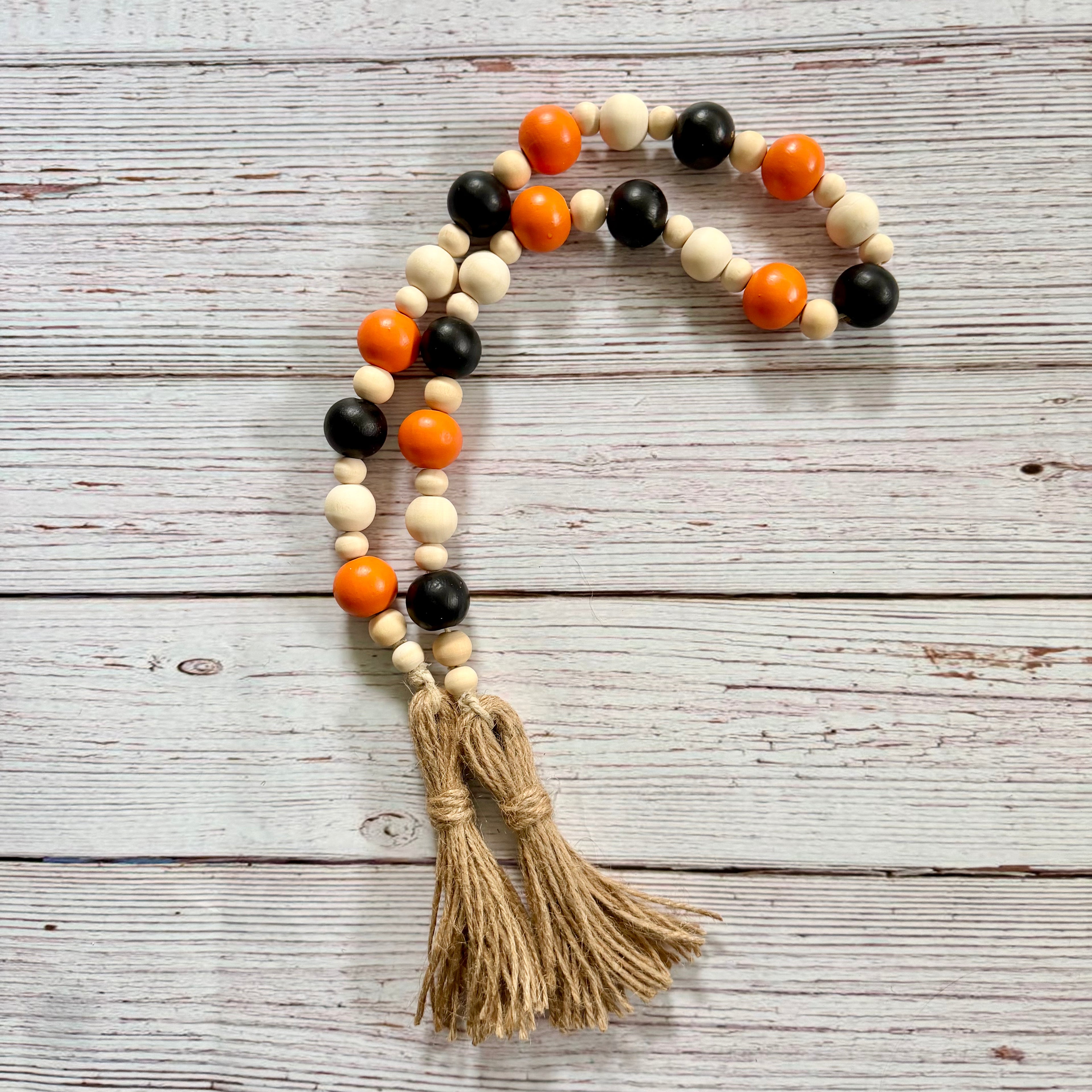A beaded garland with alternating black, cream, and orange wooden beads, finished with two knotted jute tassels, displayed on a rustic white-washed wooden background.