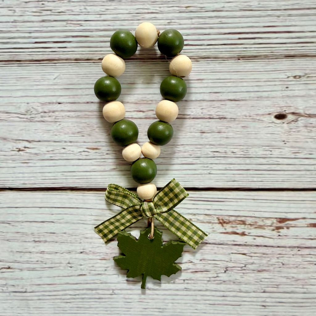 A wooden bead garland with alternating dark green and light natural wood beads, finished with a green plaid ribbon bow and a green maple leaf charm, displayed on a rustic white-washed wood background.