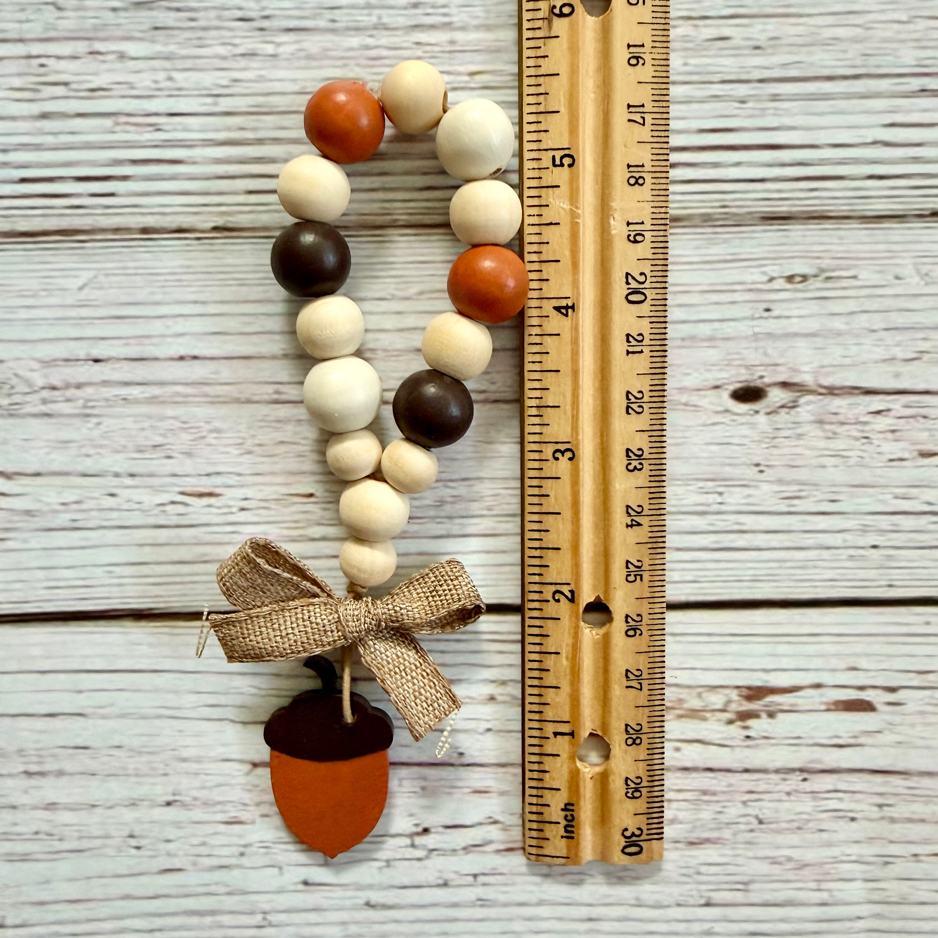 Acorn bead garland with wooden acorns in cream, orange, and dark brown, tied with a burlap bow and finished with a wooden acorn charm, shown next to a wooden ruler on a whitewashed wood background.