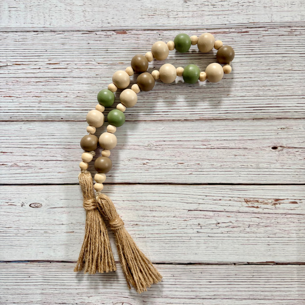 A bohemian-style wooden bead garland with natural wood beads, sage green beads, and two tan jute tassels, displayed on a rustic white-washed wooden background.