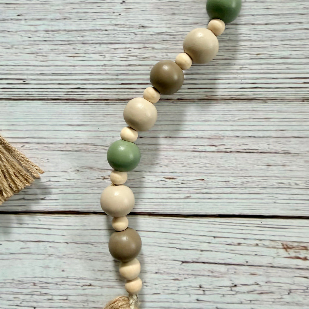 A beaded garland with alternating matte sage green, natural cream, and taupe wooden beads, laid diagonally on a distressed white wood plank surface.