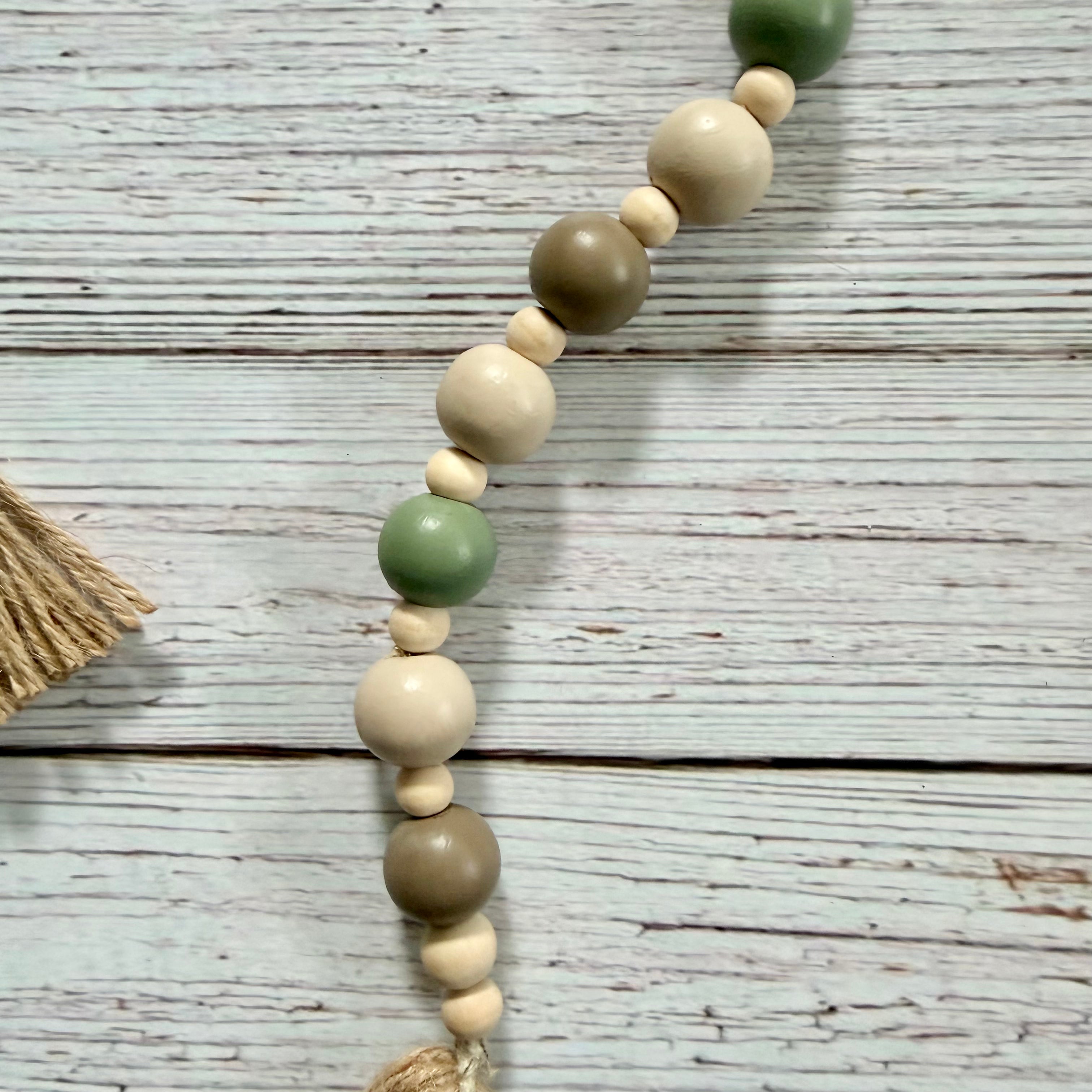 A beaded garland with alternating matte sage green, natural cream, and taupe wooden beads, laid diagonally on a distressed white wood plank surface.