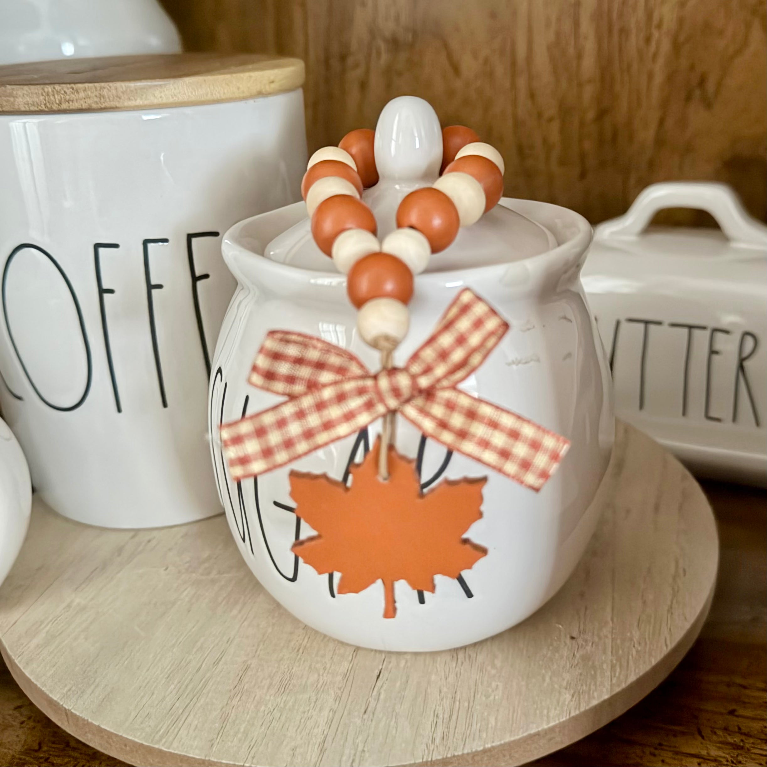 A white ceramic canister with a lid, decorated with an orange maple leaf and a red-and-white gingham ribbon bow, topped with a beaded garland. It is styled on a round wooden tray alongside other white canisters in a warm kitchen setting.