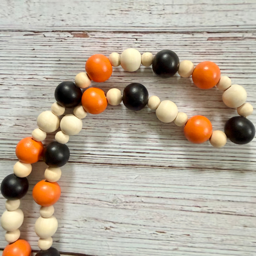 A beaded garland made of alternating matte black, natural, and bright orange round beads, arranged on a distressed white wood background.