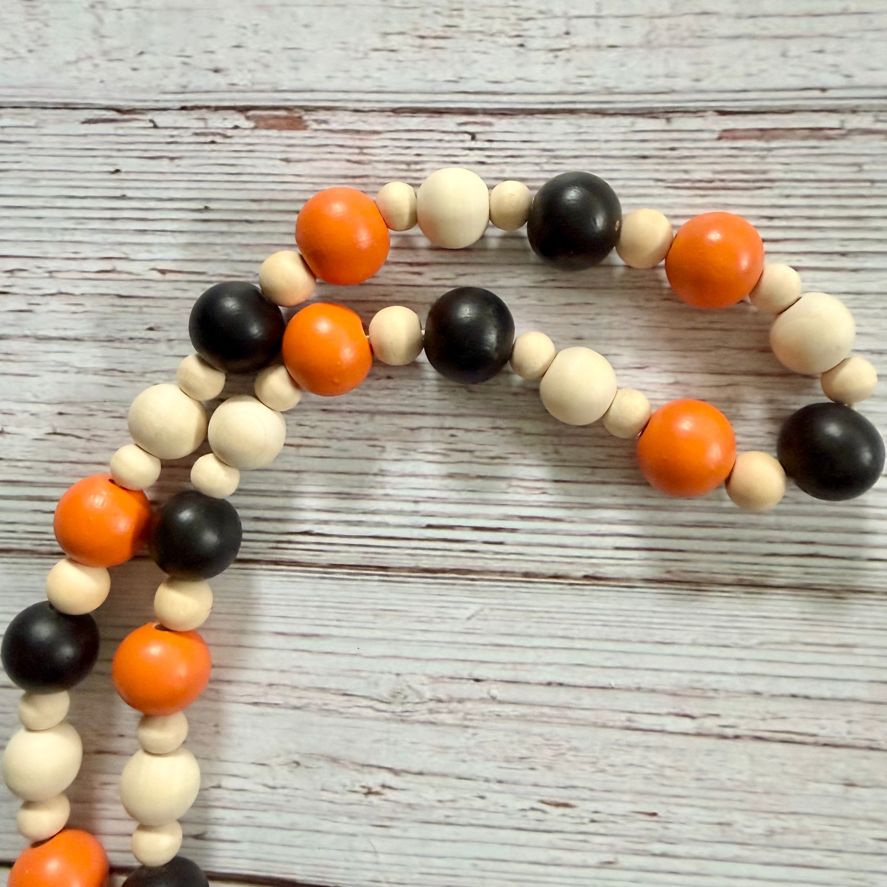 A beaded garland made of alternating matte black, natural, and bright orange round beads, arranged on a distressed white wood background.