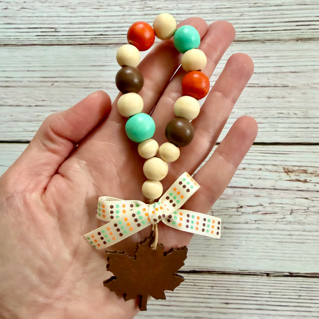 A hand holds a beaded autumn-themed charm with orange, teal, brown, and cream wooden beads, a patterned ribbon bow, and a brown leaf-shaped ornament against a whitewashed wood background.