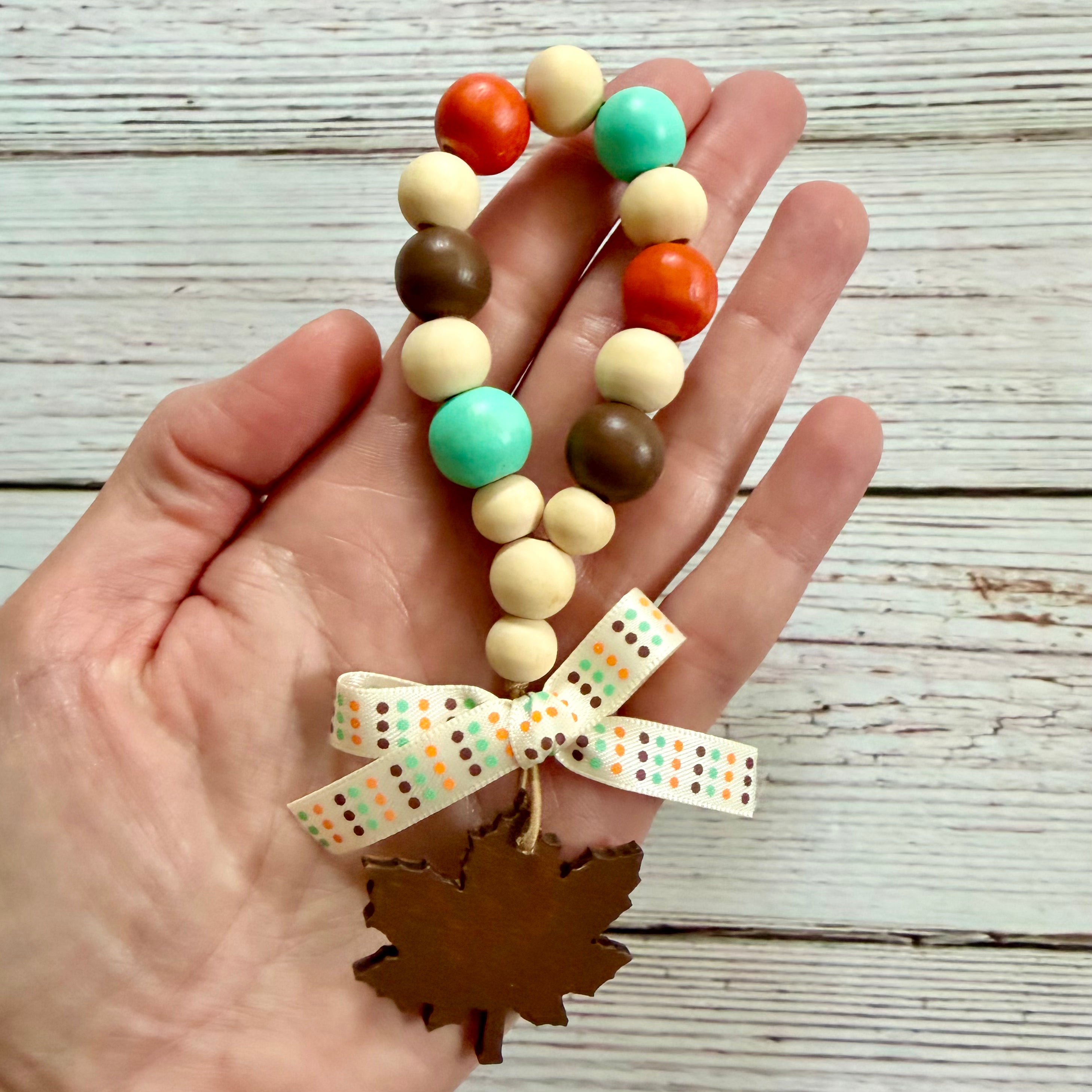 A hand holds a beaded autumn-themed charm with orange, teal, brown, and cream wooden beads, a patterned ribbon bow, and a brown leaf-shaped ornament against a whitewashed wood background.