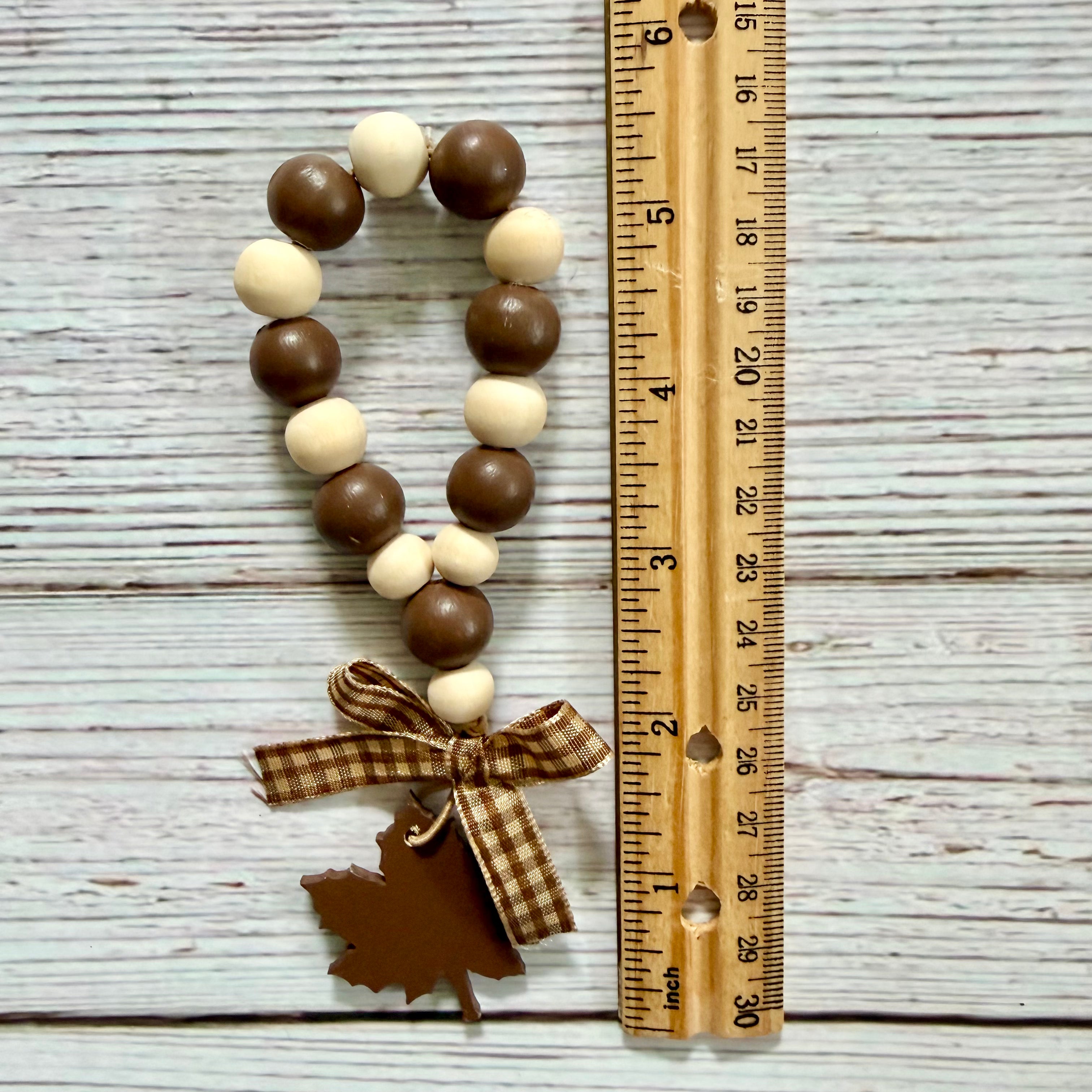 A beaded ornament or keychain charm made of alternating brown and cream wooden beads, tied with a brown gingham ribbon and finished with a small brown leaf-shaped tag, shown next to a wooden ruler on a whitewashed wood background.