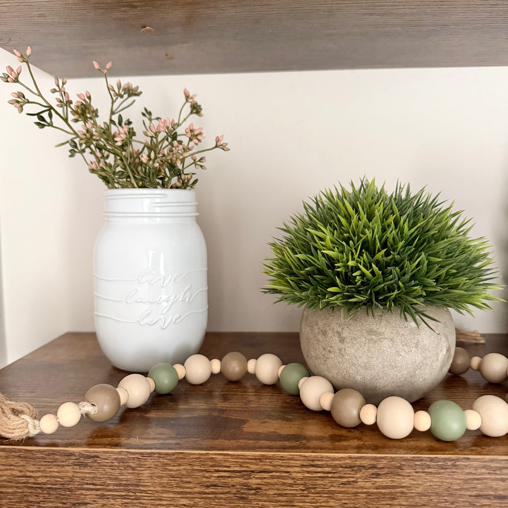 A styled rustic shelf display featuring a white mason jar with dried pink flowers, a green artificial grass plant in a concrete pot, and a wooden bead garland draped across the shelf.