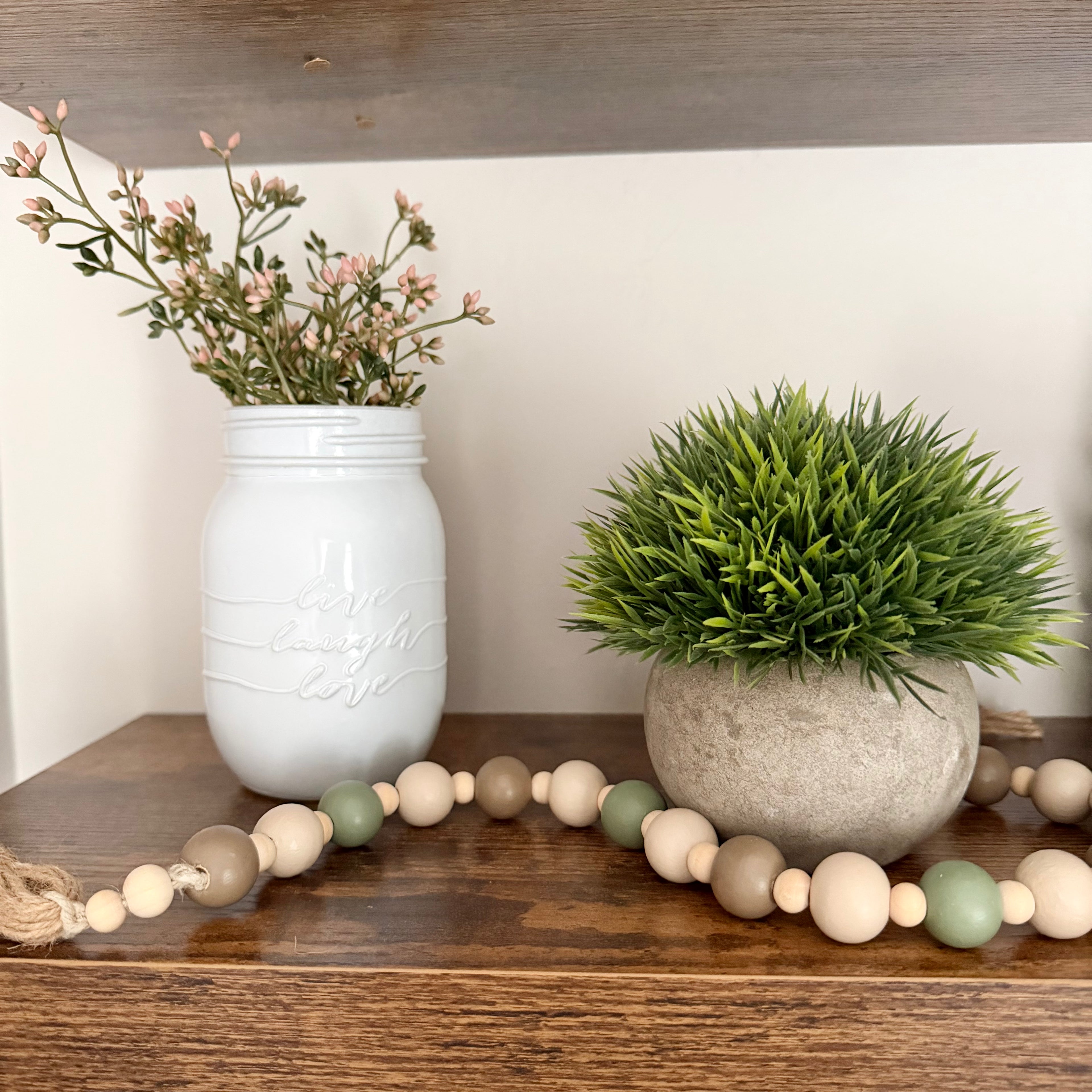 A styled rustic shelf display featuring a white mason jar with dried pink flowers, a green artificial grass plant in a concrete pot, and a wooden bead garland draped across the shelf.