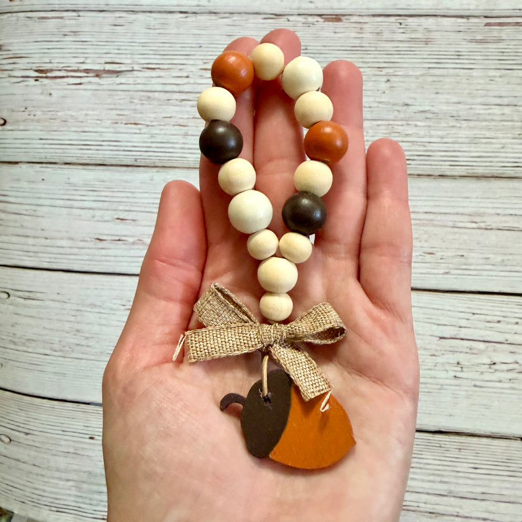 A hand holding a beaded autumn-themed charm bracelet with orange, cream, and dark brown wooden beads, a burlap ribbon bow, and a wooden acorn and pumpkin charm, set against a rustic white-washed wood plank background.