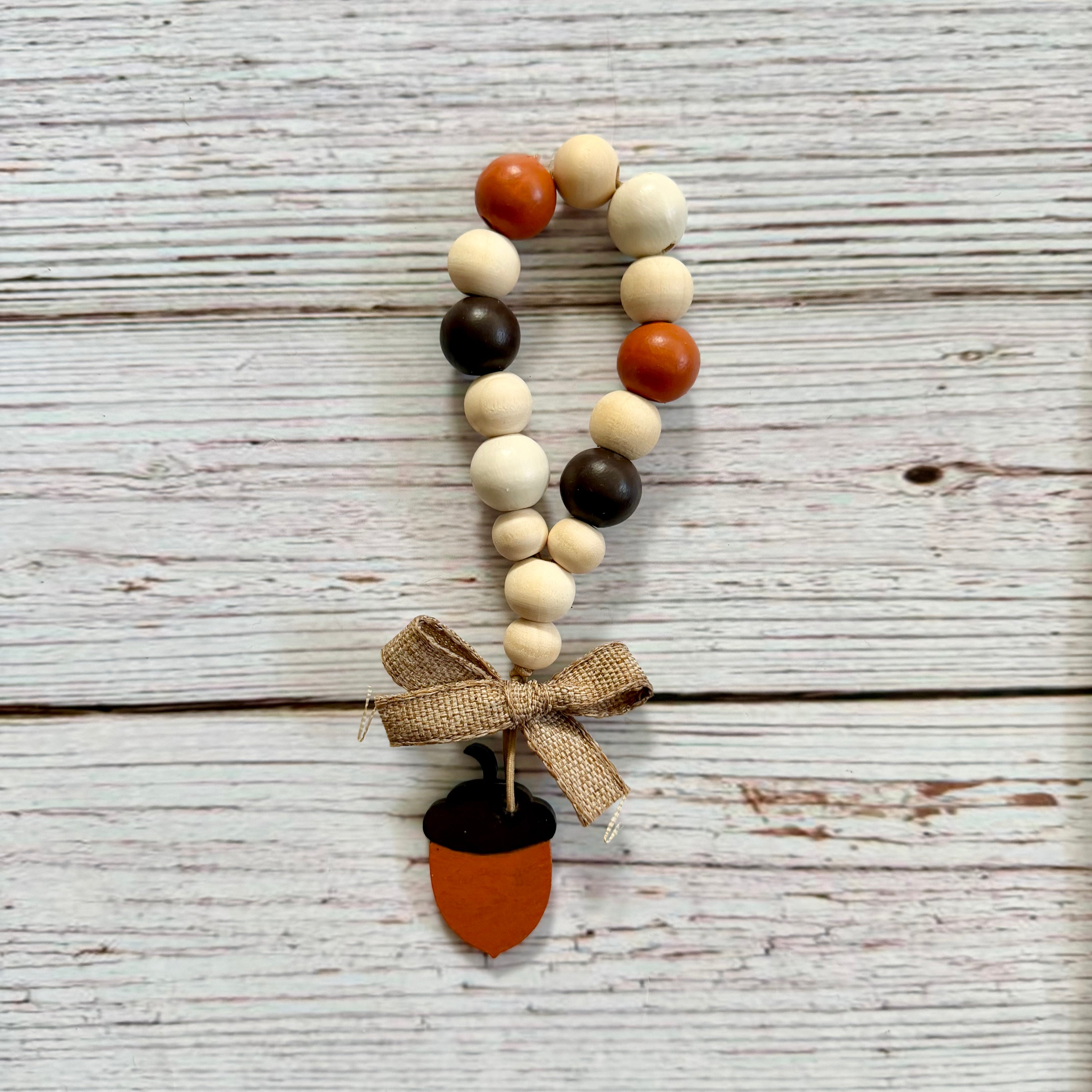 Autumn-themed wooden bead garland with orange, cream, and dark brown beads, tied with a burlap ribbon and finished with an acorn charm, displayed on a rustic white-washed wood background.