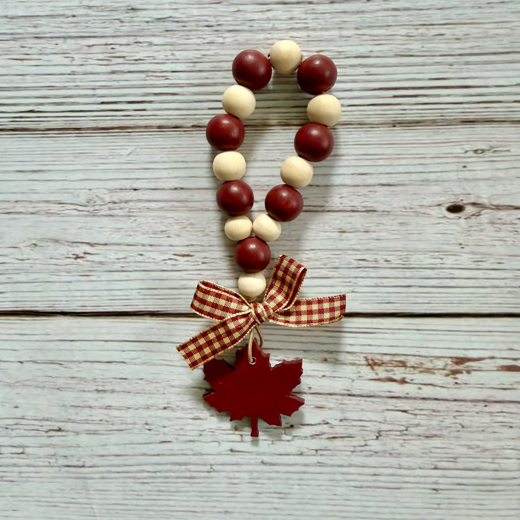A beaded garland with alternating dark red and cream wooden beads, finished with a red-and-white gingham ribbon bow and a red maple leaf charm, displayed on a whitewashed wood background.