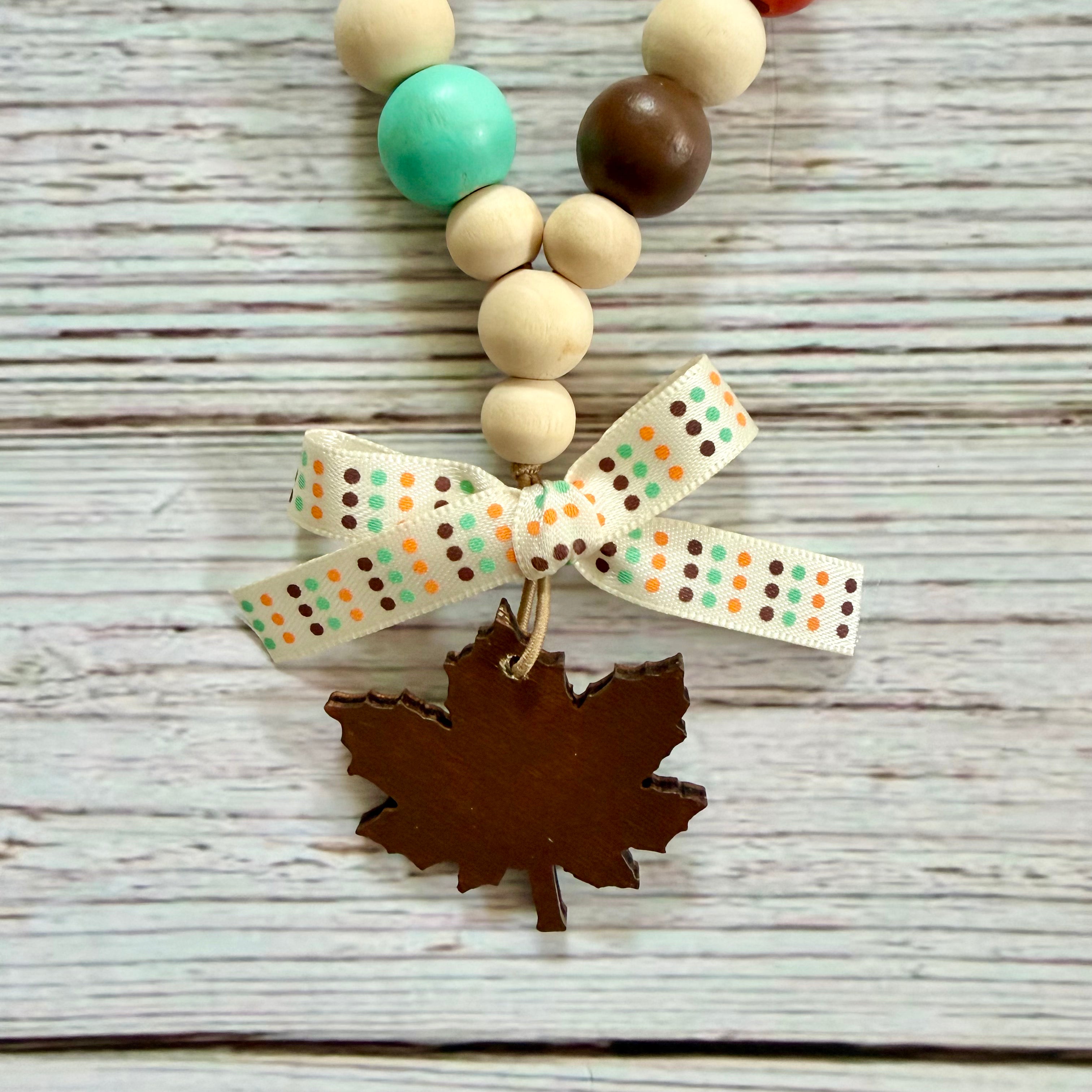 A beaded necklace with natural wood beads, a turquoise bead, a dark brown bead, and a patterned white ribbon bow tied above a dark brown maple leaf charm, displayed on a whitewashed wood background.