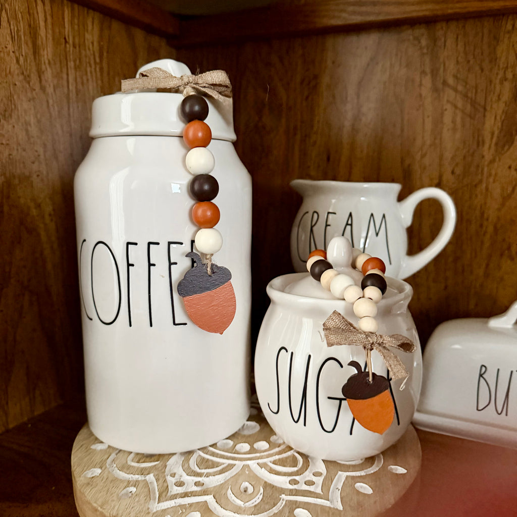 White ceramic canister and sugar bowl canisters with black lettering, decorated with acorn charms and wooden bead garlands, displayed on a wooden shelf.