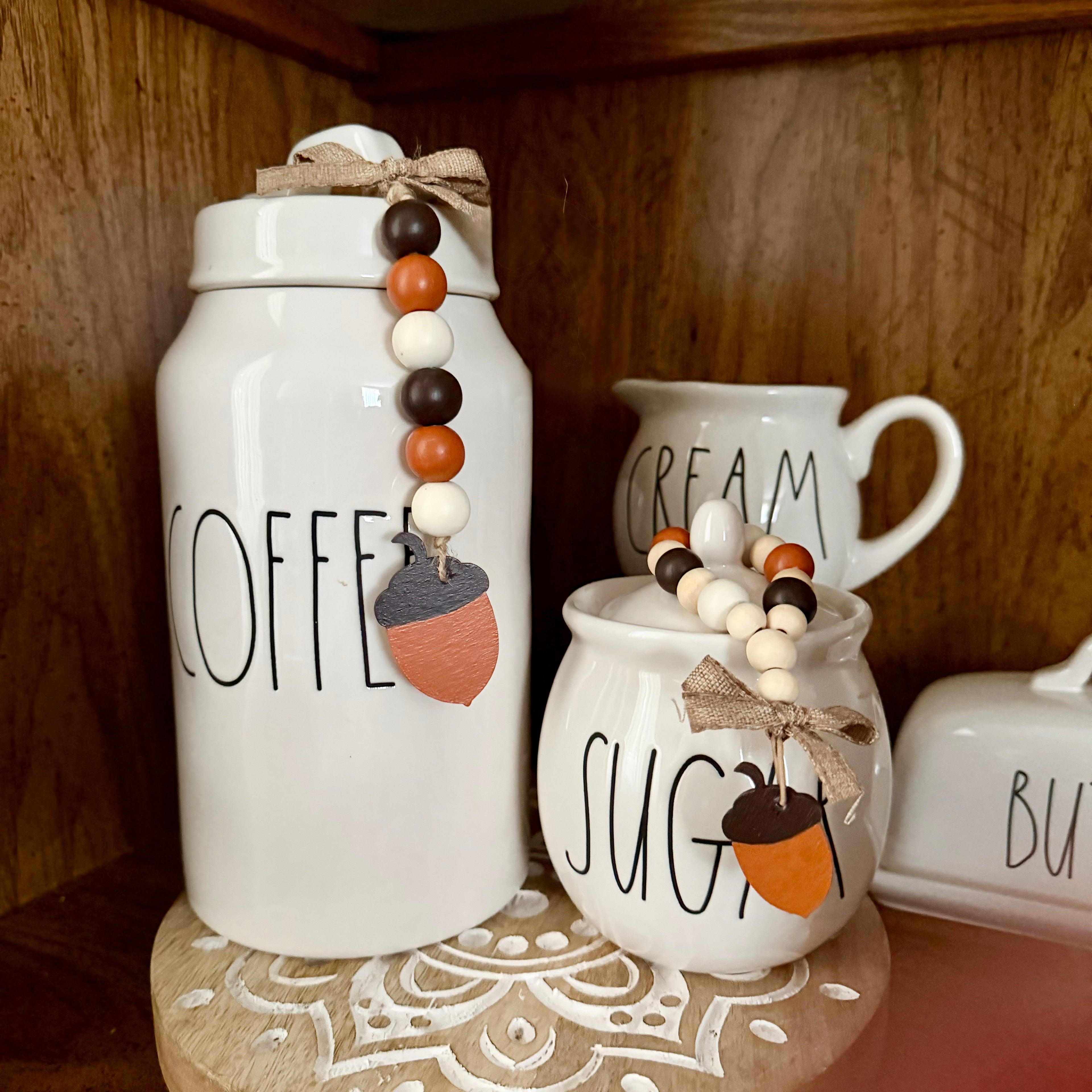 White ceramic canister and sugar bowl canisters with black lettering, decorated with acorn charms and wooden bead garlands, displayed on a wooden shelf.