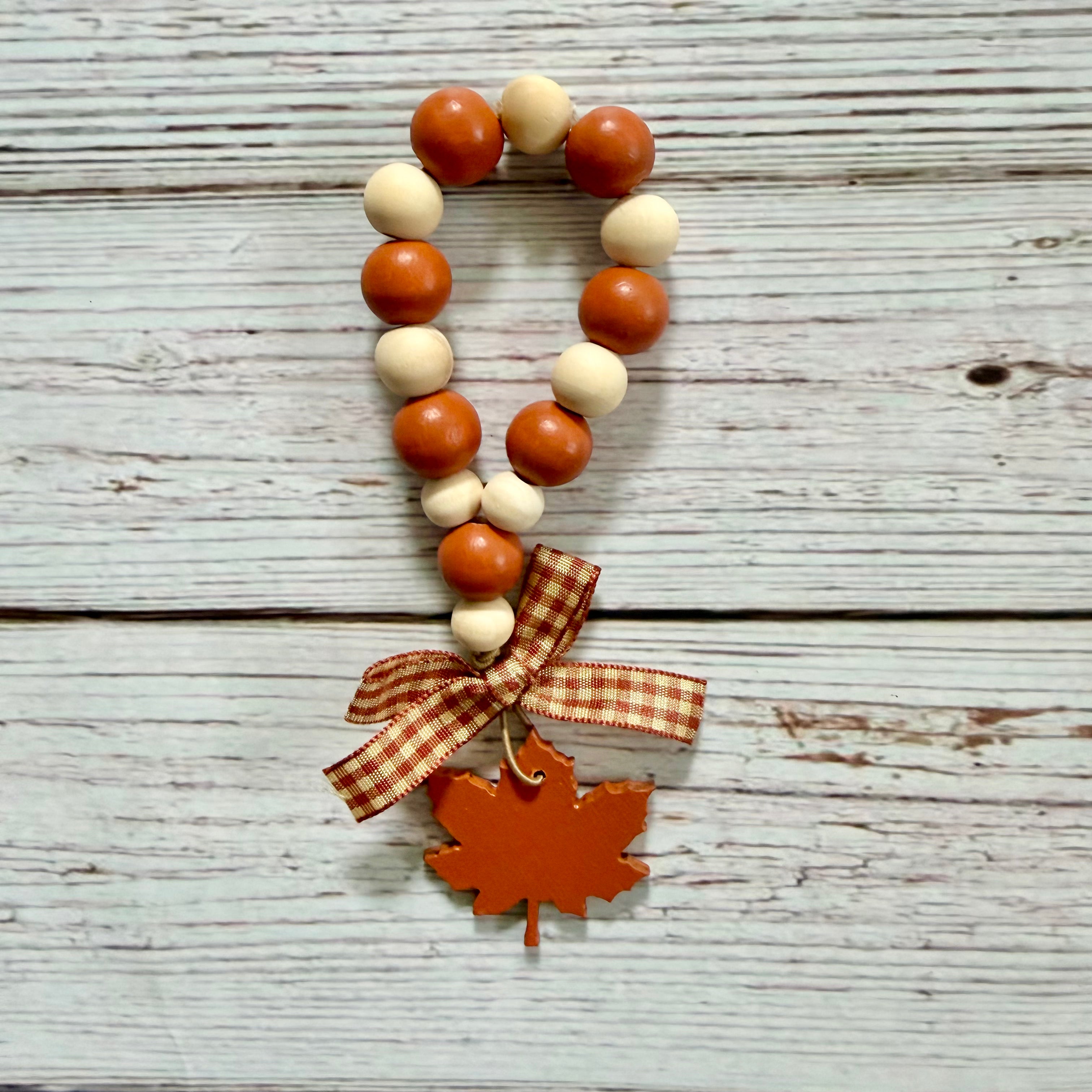 Autumn-themed wooden bead garland with orange and cream beads, a gingham ribbon bow, and a wooden maple leaf charm, displayed on a rustic white-washed wood background.