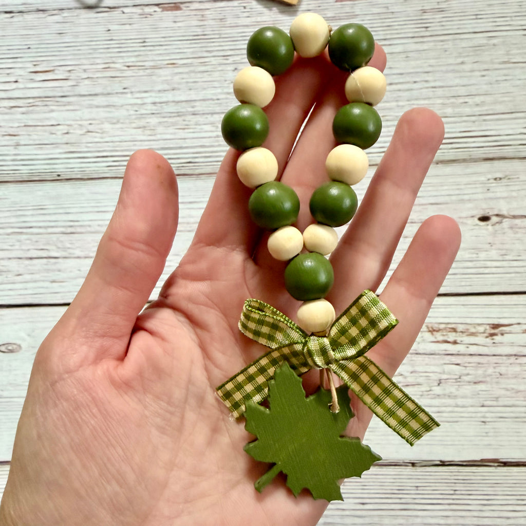 A hand holding a beaded garland made of green and cream wooden beads, tied with a green and beige gingham ribbon and finished with a green leaf charm, set against a rustic white-washed wood background.