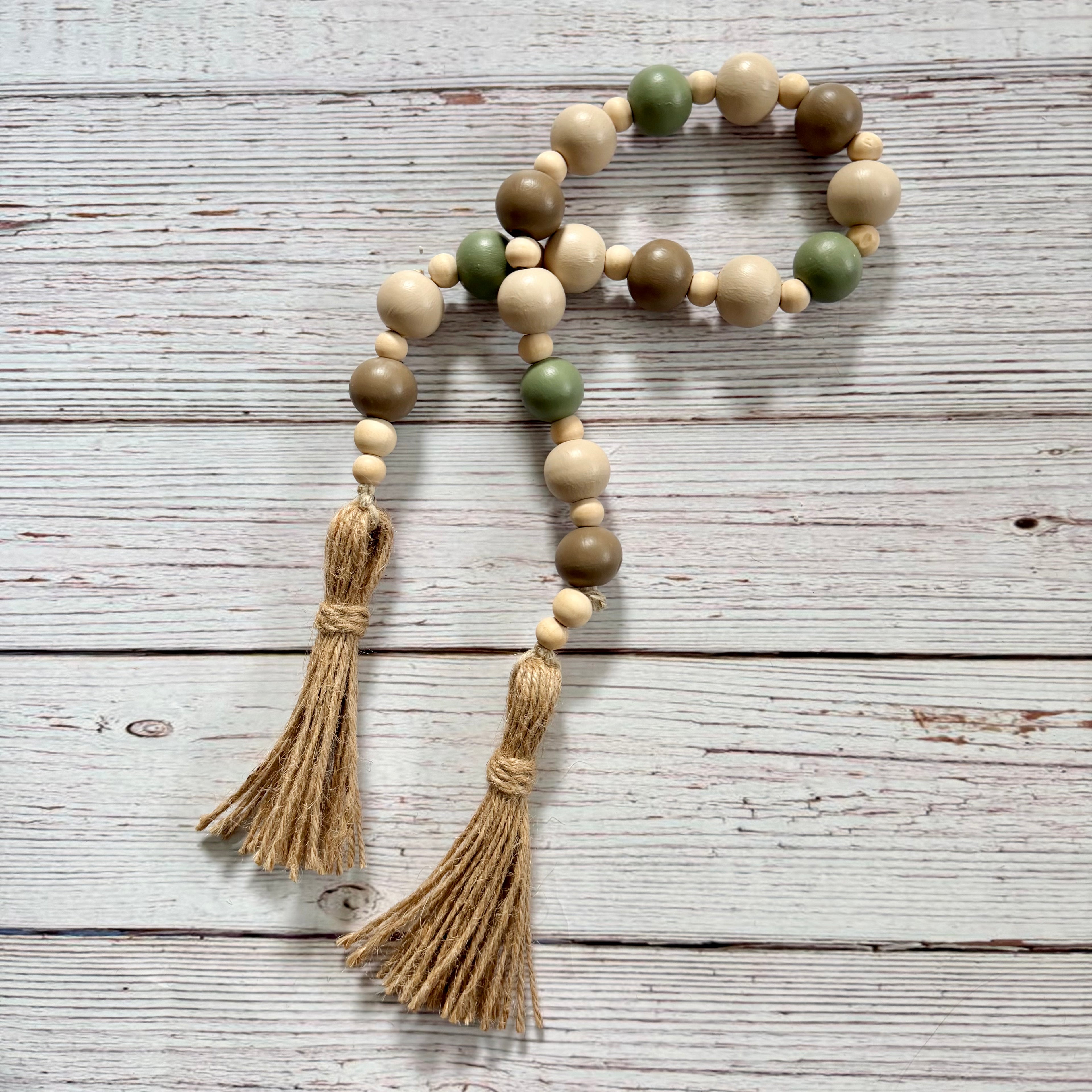 A beaded garland with natural wood beads, sage green and taupe accent beads, and two long jute tassels, laid flat on a whitewashed wooden surface.