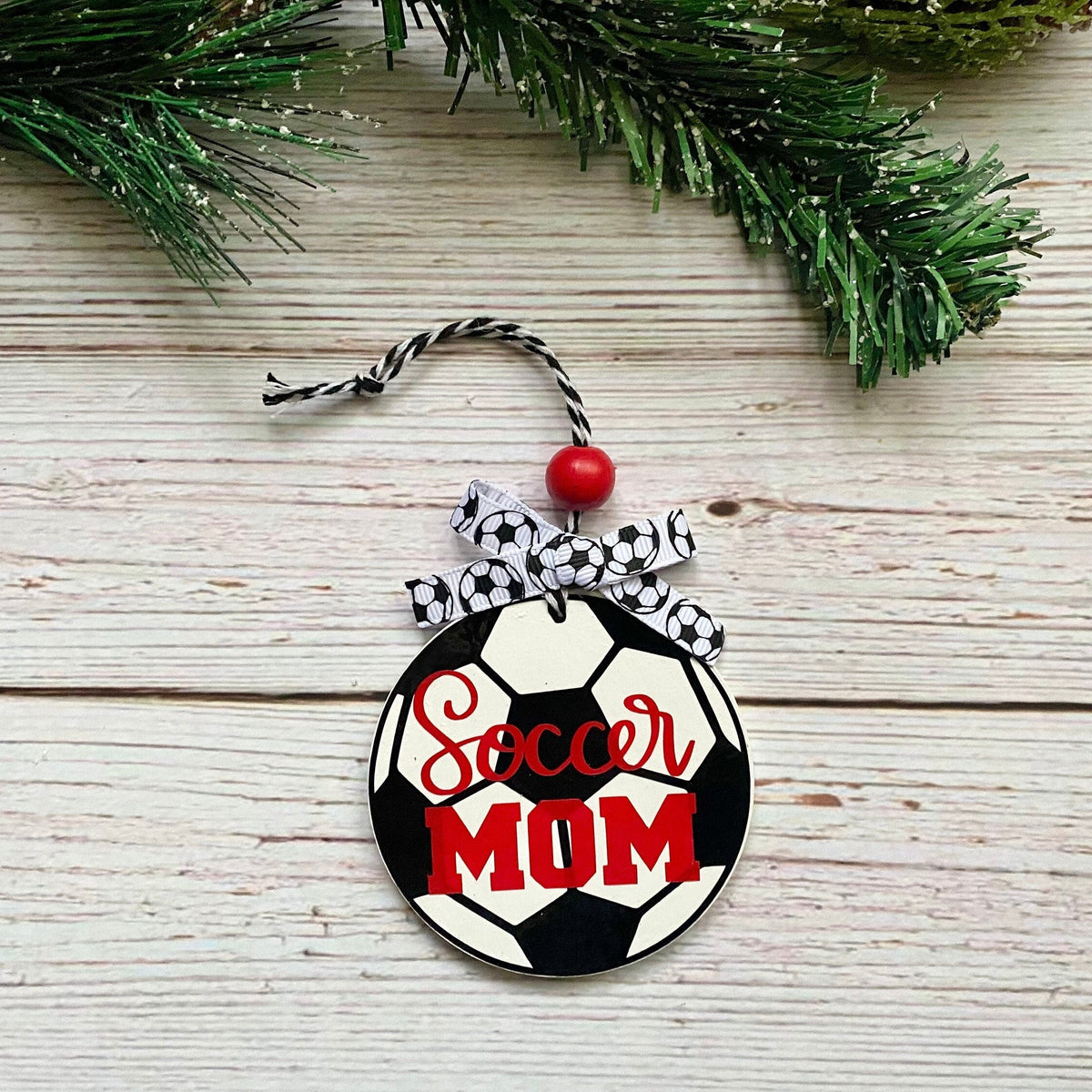 A round wooden ornament shaped like a soccer ball with the words 'Soccer Mom' in red script and block lettering, hanging from a black-and-white patterned ribbon and a red bead on a whitewashed wood surface with green pine branches at the top.