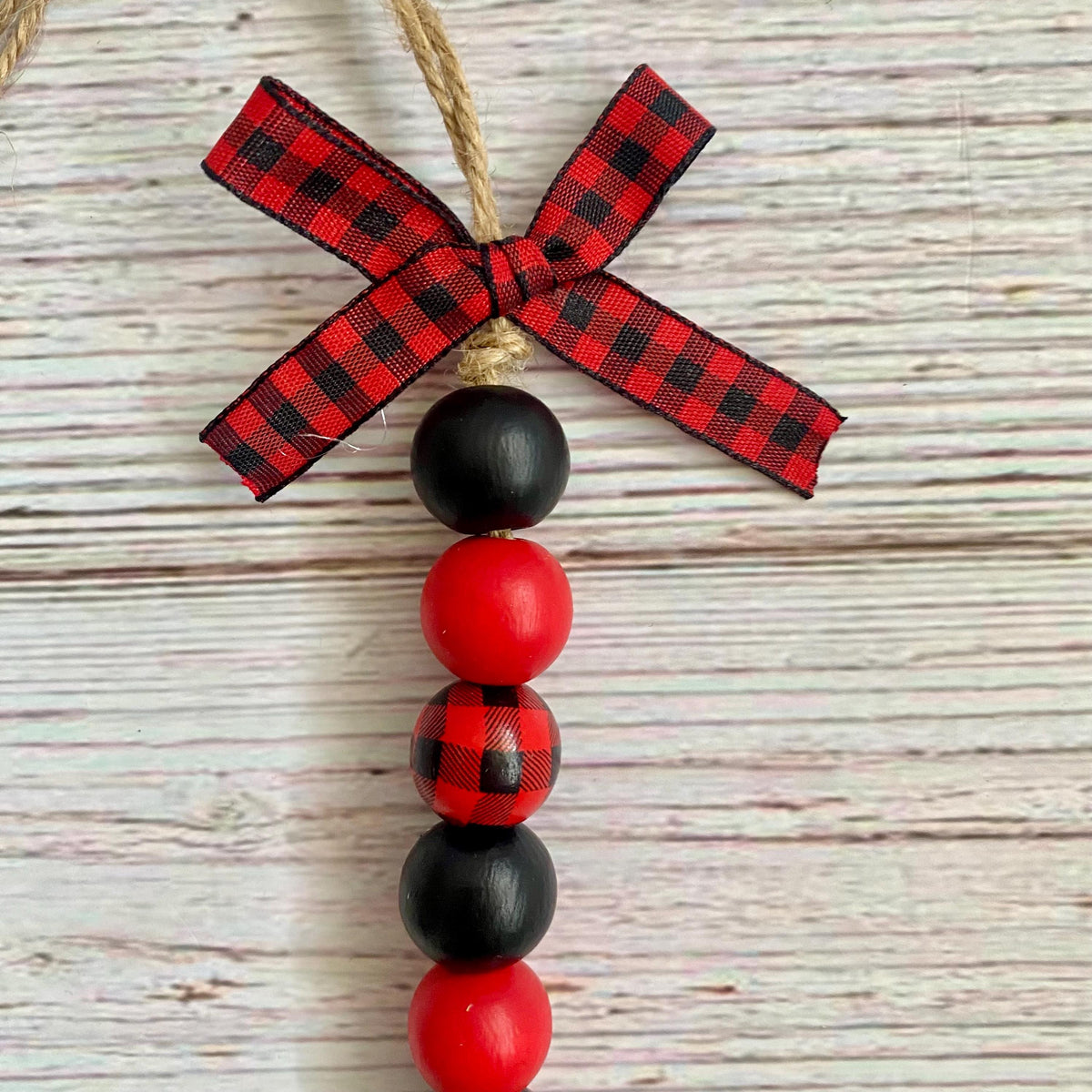 A rustic Christmas ornament featuring a red and black buffalo plaid ribbon bow at the top, hanging from a natural jute string. Below the bow is a vertical sequence of wooden beads: one solid black, one red, one plaid, one solid black, and one solid red, all resting on a whitewashed wood plank background.