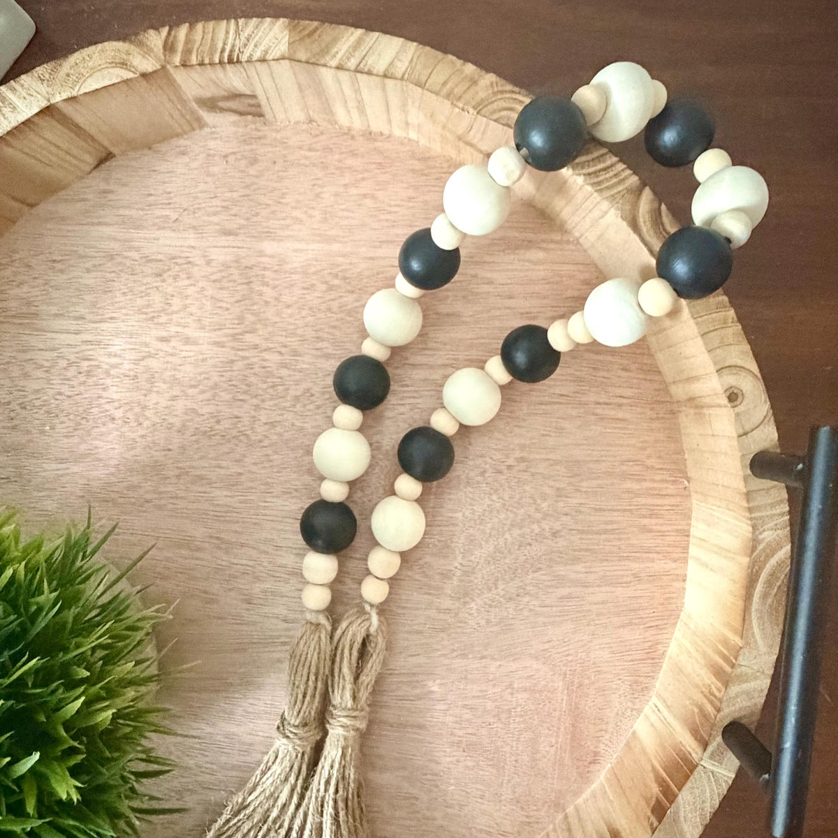 A beaded garland with alternating matte black and natural cream wooden beads, finished with two knotted jute tassels, displayed on a round wooden serving tray.