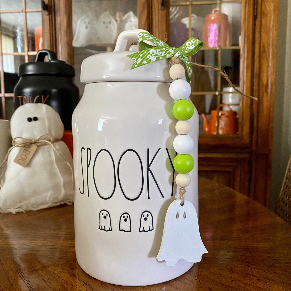 A white ceramic Halloween cookie jar with 'SPOOKY' and ghost illustrations on the front, decorated with a green patterned ribbon, wooden beads, and a white ghost charm, sitting on a wooden table.