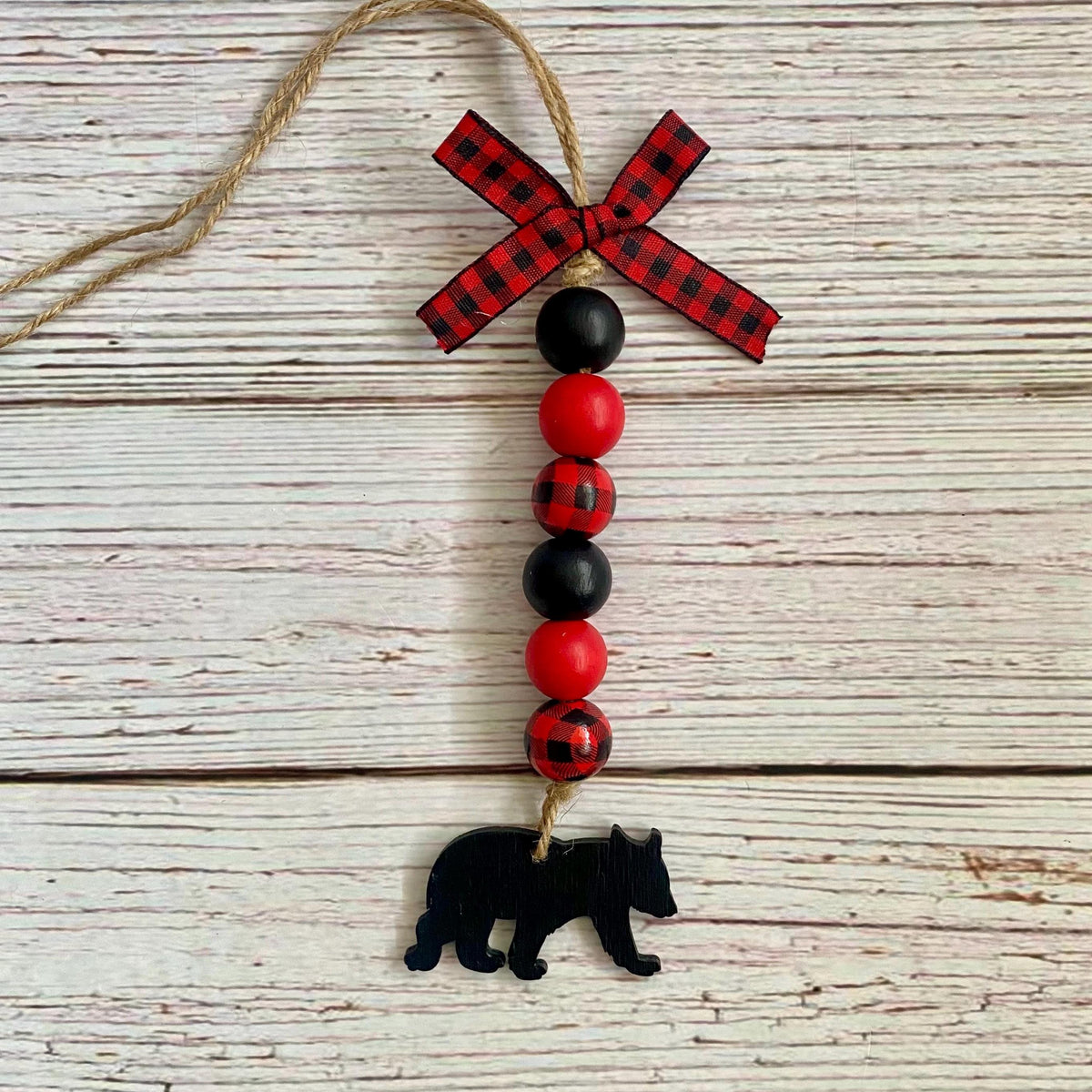 A rustic Christmas ornament featuring red and black beads, a red and black buffalo plaid ribbon bow at the top, and a black bear silhouette charm at the bottom, hanging from a jute twine string against a whitewashed wood plank background.