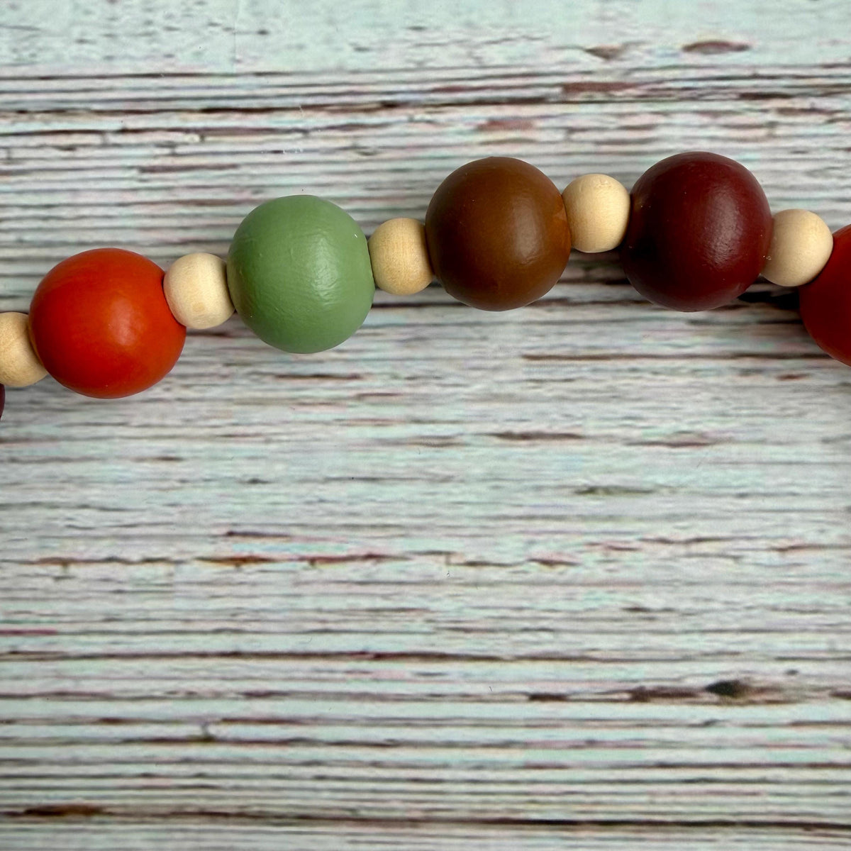 A close-up of a wooden bead garland with large and small painted beads in red, green, brown, and dark red, strung on a black cord over a distressed white wood background.