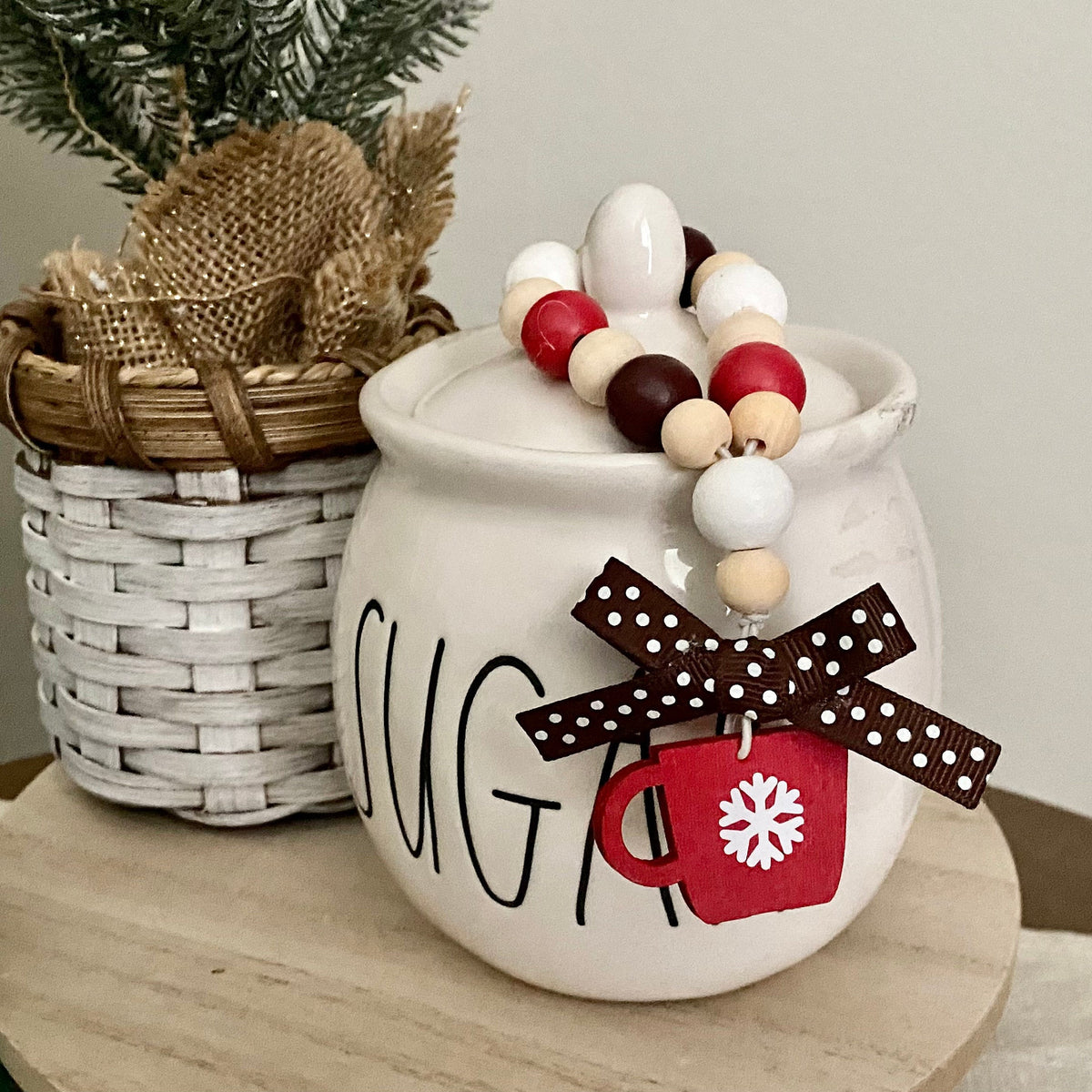 A white ceramic sugar bowl with 'SUGAR' written on the front, decorated with a beaded garland and a red mug charm with a brown polka-dot ribbon. It is styled on a wooden stand next to a white woven basket with burlap and evergreen branches.