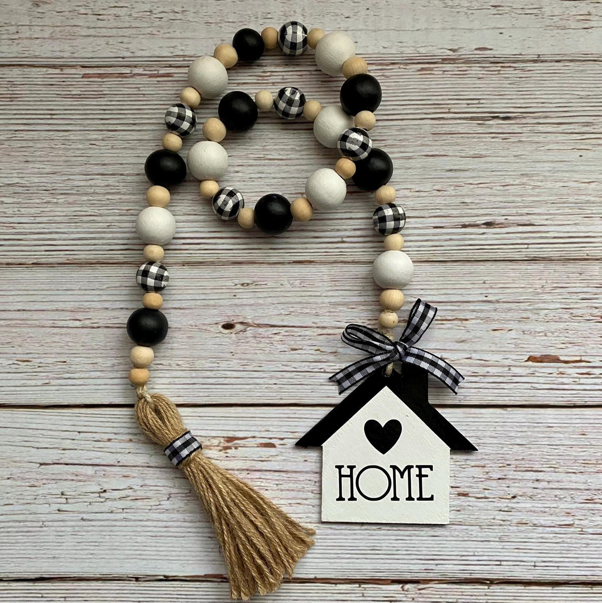 A rustic wooden bead garland with black, white, and gingham pattern beads, a jute tassel, and a white and black house-shaped ornament that says HOME, displayed on a whitewashed wood background.