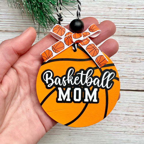 A hand holding a round basketball-themed Christmas ornament with the words 'Basketball MOM' in white script and block lettering. The ornament is orange with black basketball-style lines, decorated with an orange-and-white patterned ribbon bow at the top, and shown against a light wood plank background with a small green pine branch at the top.