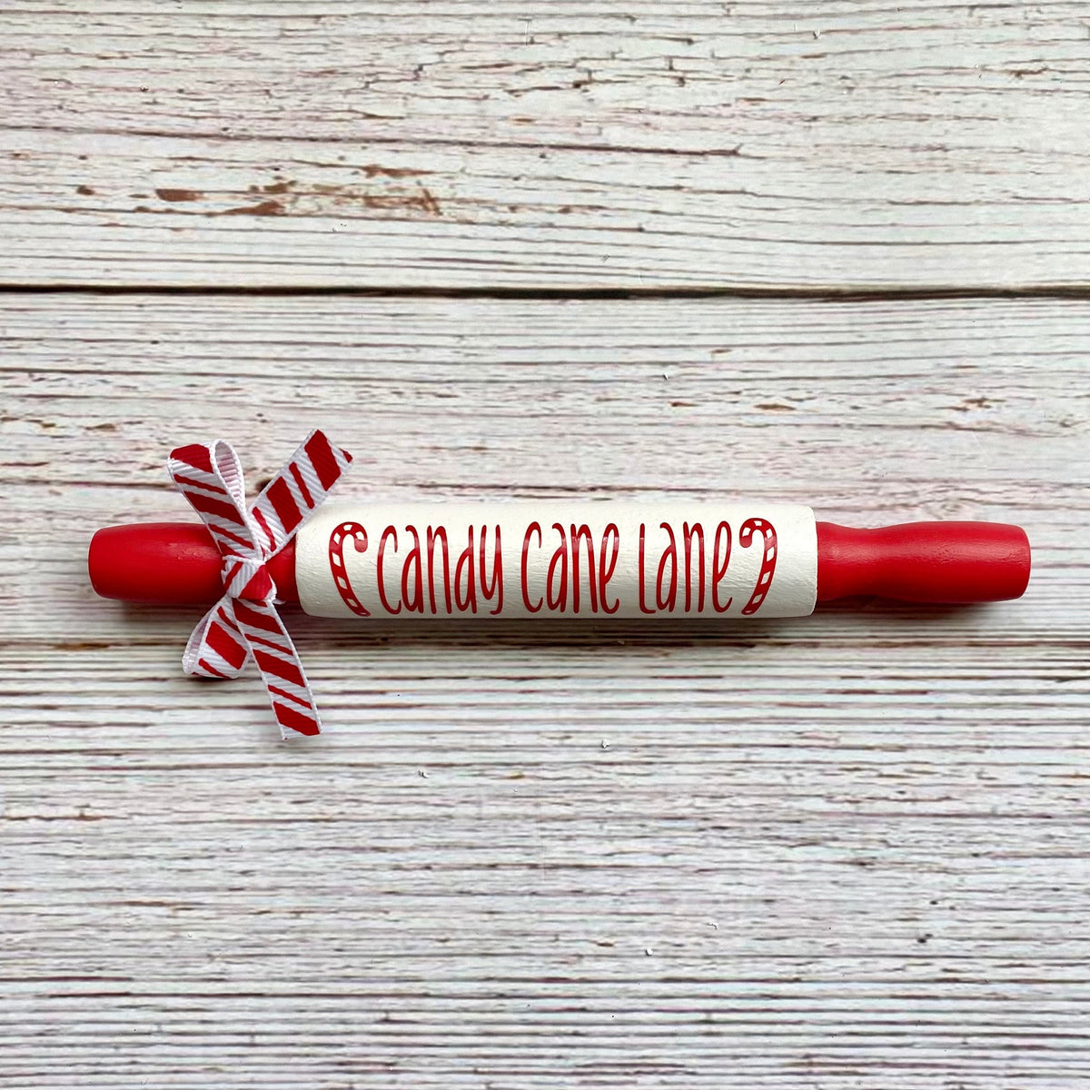 A red and white wooden rolling pin with 'candy cane lane' written on the center in red script, tied with a red and white striped ribbon, on a rustic white wood background.