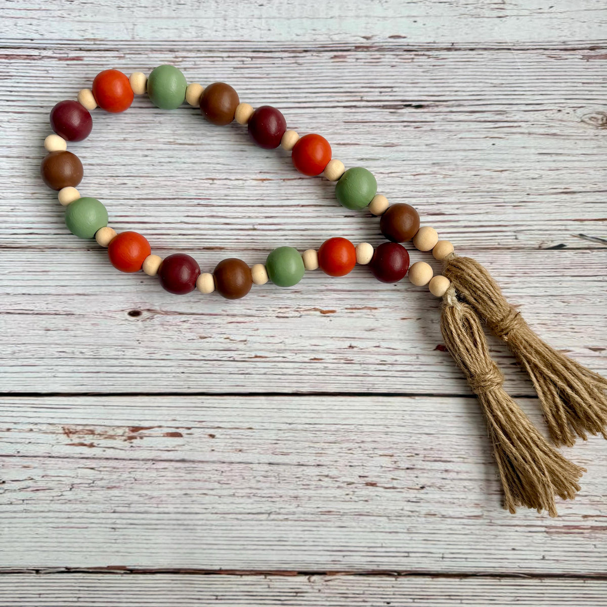 A beaded garland with wooden beads in red, green, brown, and natural beige, featuring a natural jute tassel, displayed on a rustic white-washed wooden background.