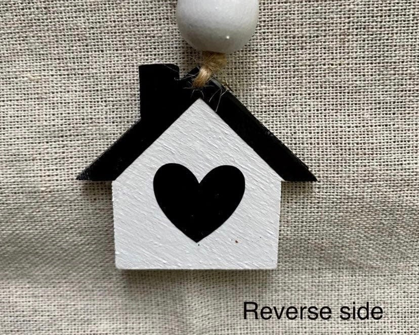 A small house-shaped ornament or charm with a white body, black roof, and black heart in the center, hanging from a white bead on a textured beige fabric background. Text in the lower right says 'Reverse side'.